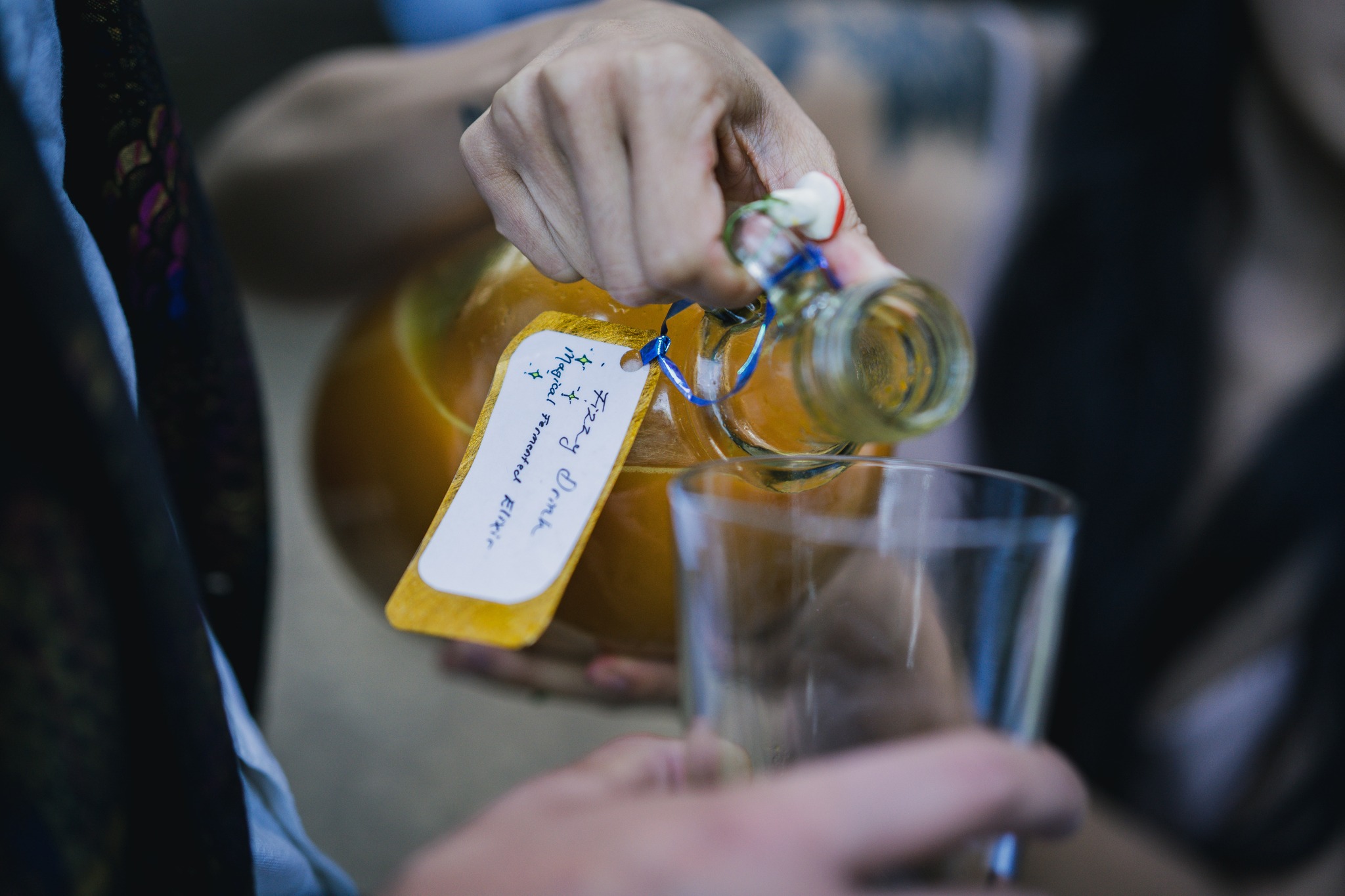 A hand is pouring a yellowish liquid from a glass bottle with a handwritten tag into a clear glass. The tag reads &quot;Fizzy Drink, Home-made Fermented Elixir.&quot;