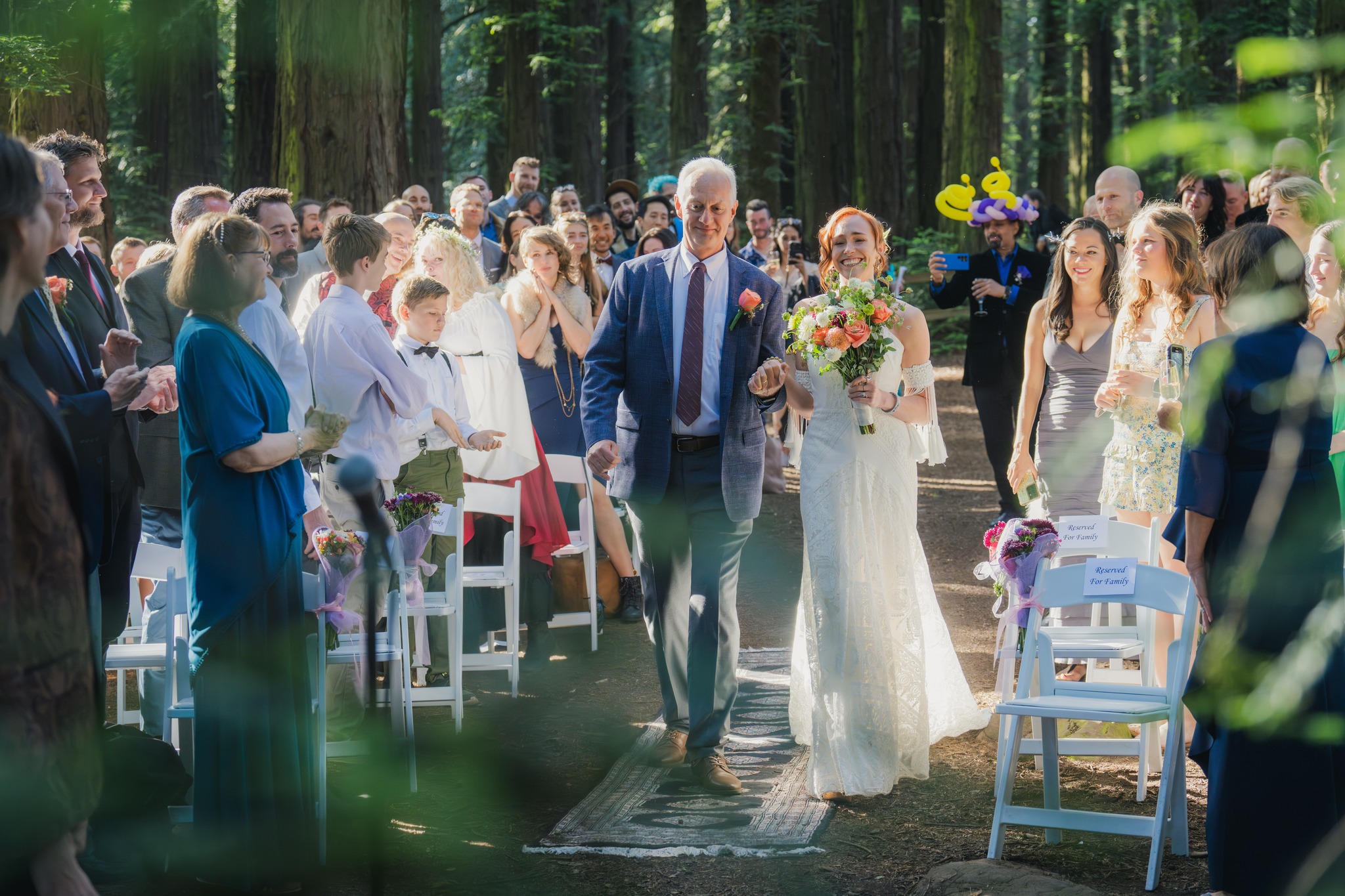 A bride in a white dress holding a bouquet walks down an outdoor aisle with an older man, surrounded by standing guests in a wooded area.