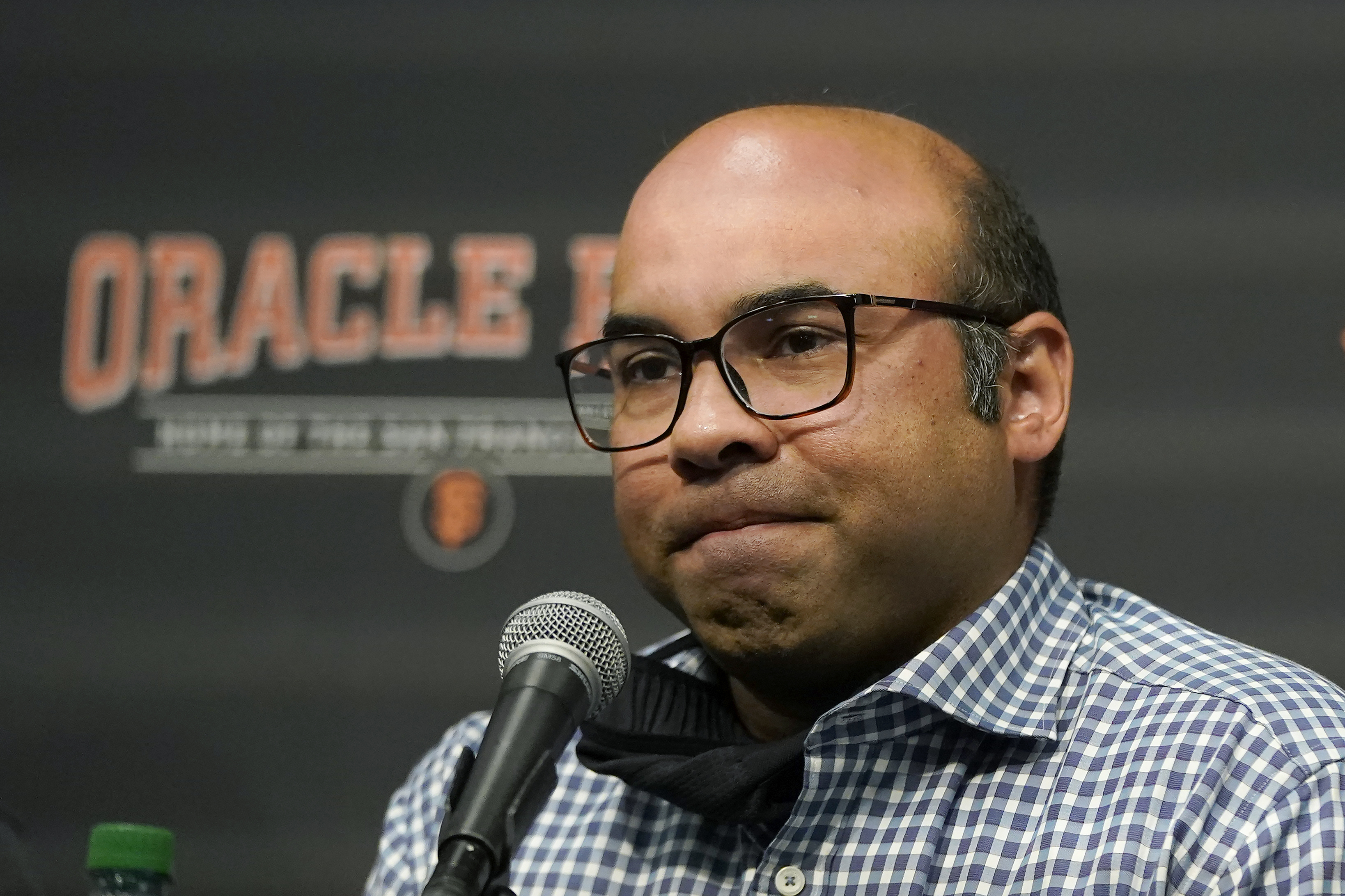 A man wearing glasses and a checkered shirt sits in front of a microphone, visibly pensive, with a dark background showing the words "Oracle Park."