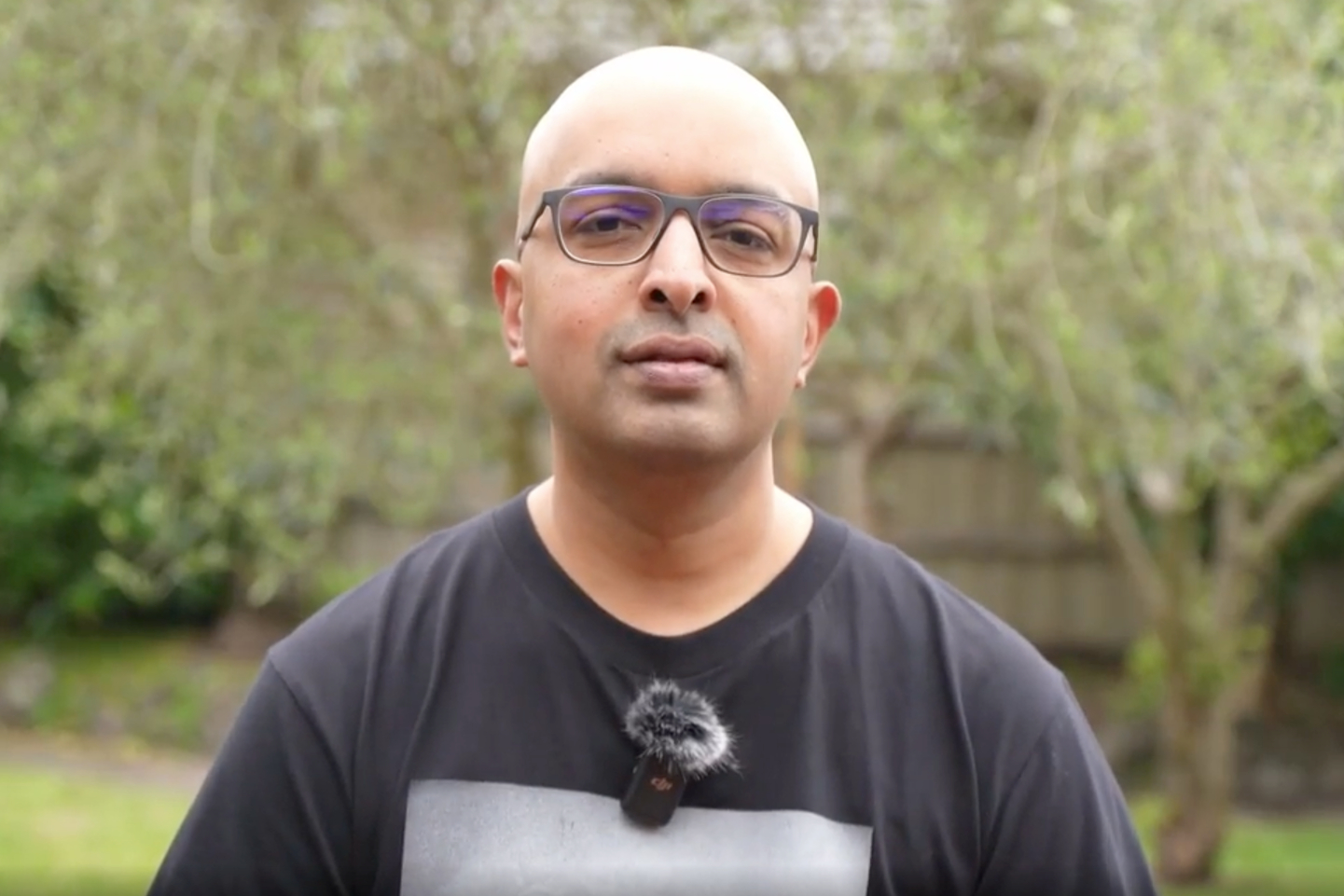 A bald man wearing glasses and a dark t-shirt is standing outdoors. He has a small microphone clipped to his shirt, and there's a blurred green garden in the background.