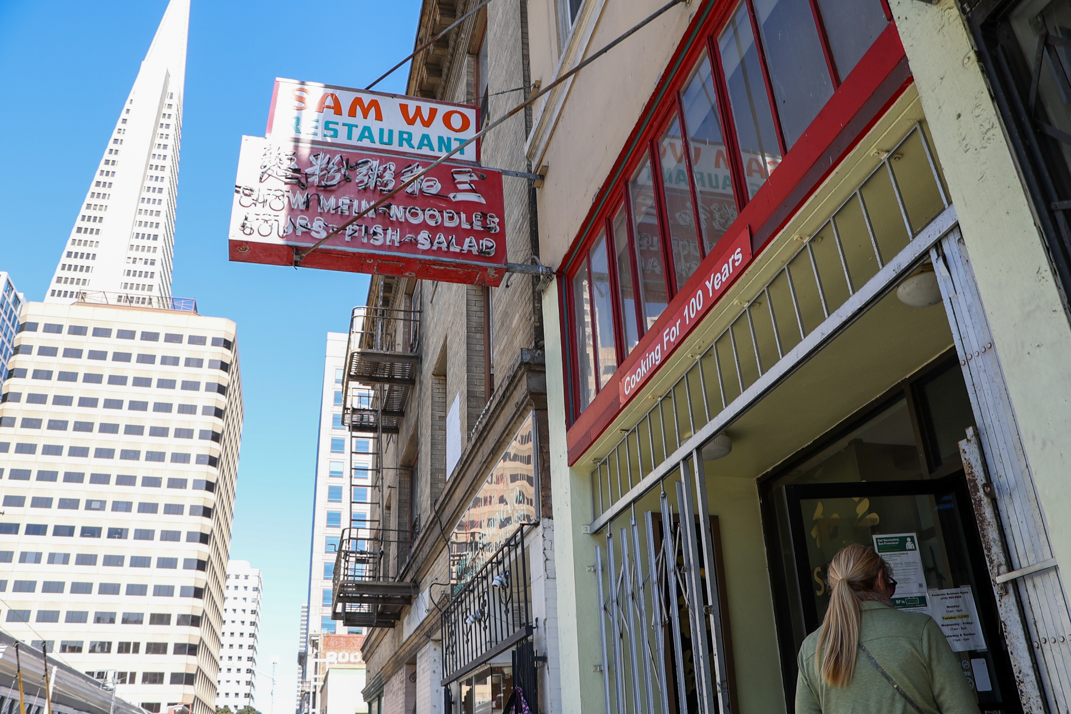 A street scene with a person entering "Sam Wo Restaurant," under a sign stating "Cooking for 100 Years." Skyscrapers, including a triangular one, are visible in the background.