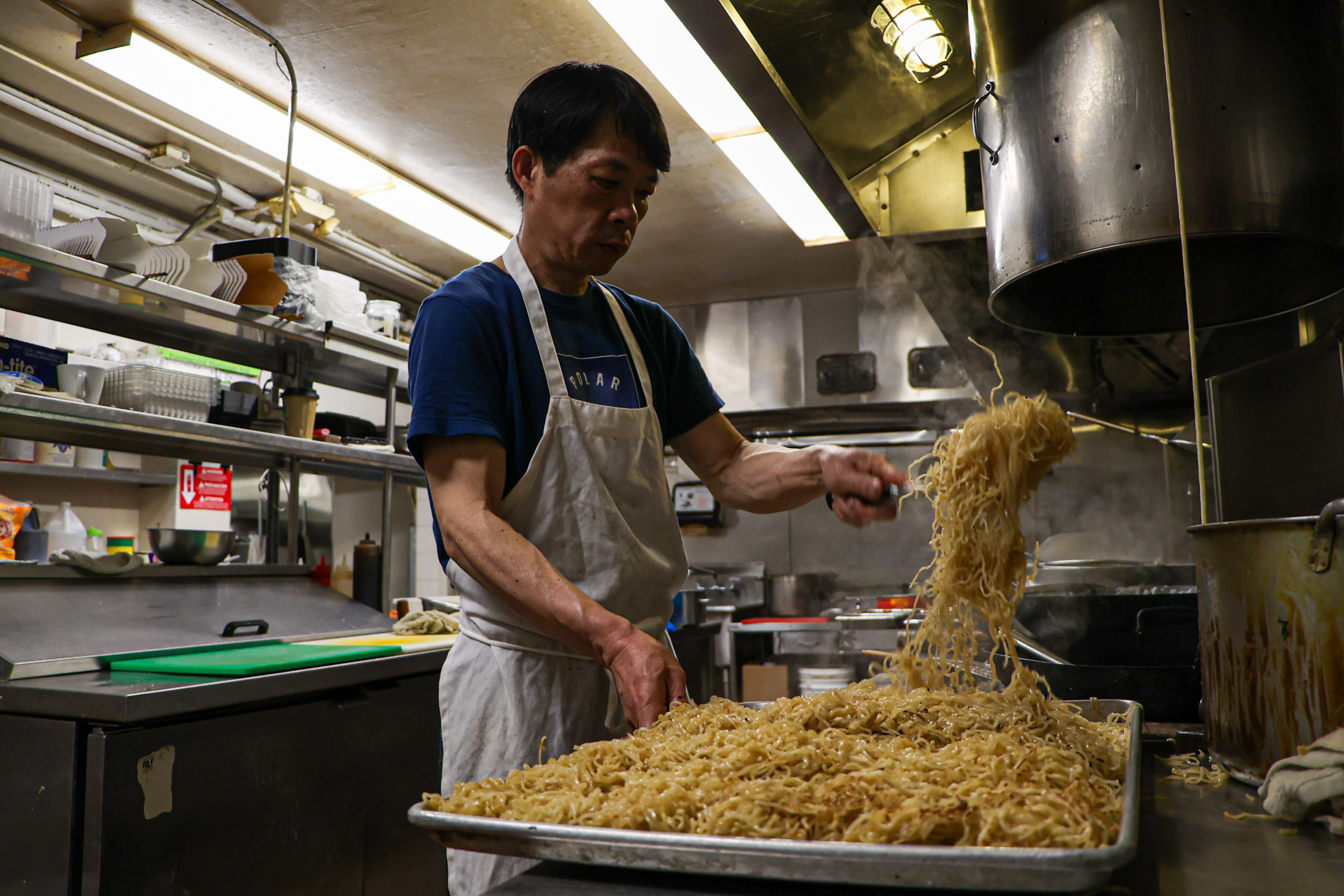 A man in a kitchen, wearing a blue shirt and white apron, is preparing a large tray of noodles. The kitchen is equipped with cooking utensils and shelves.