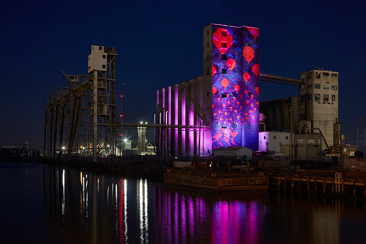 The image shows an industrial building at night, illuminated with vibrant purple and red lights, reflecting off nearby water, creating a striking visual effect.