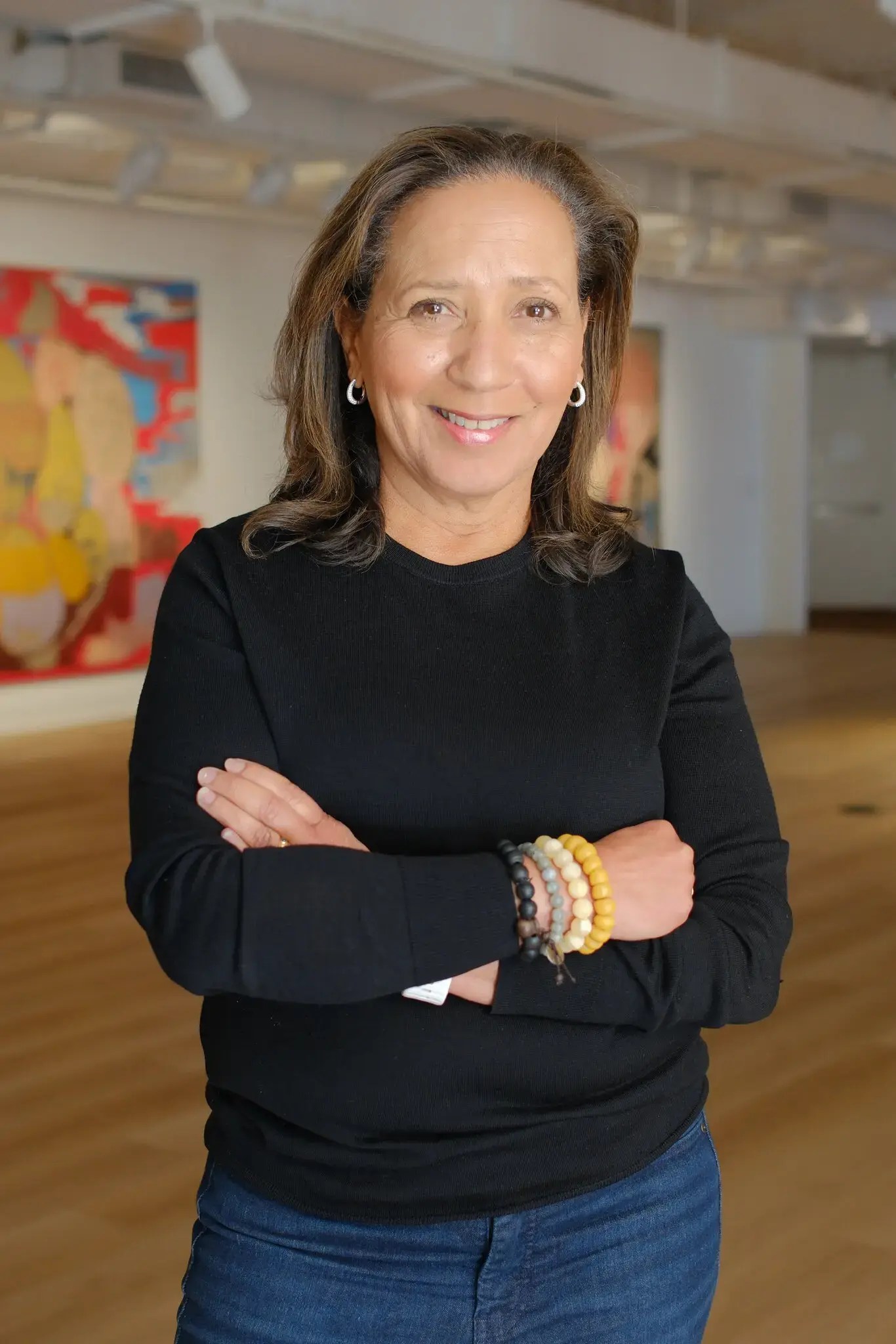 A smiling woman stands with her arms crossed, wearing a black long-sleeve shirt and blue jeans. She has shoulder-length brown hair, wears earrings, and bracelets on her wrist. The background features abstract art and a well-lit spacious room.