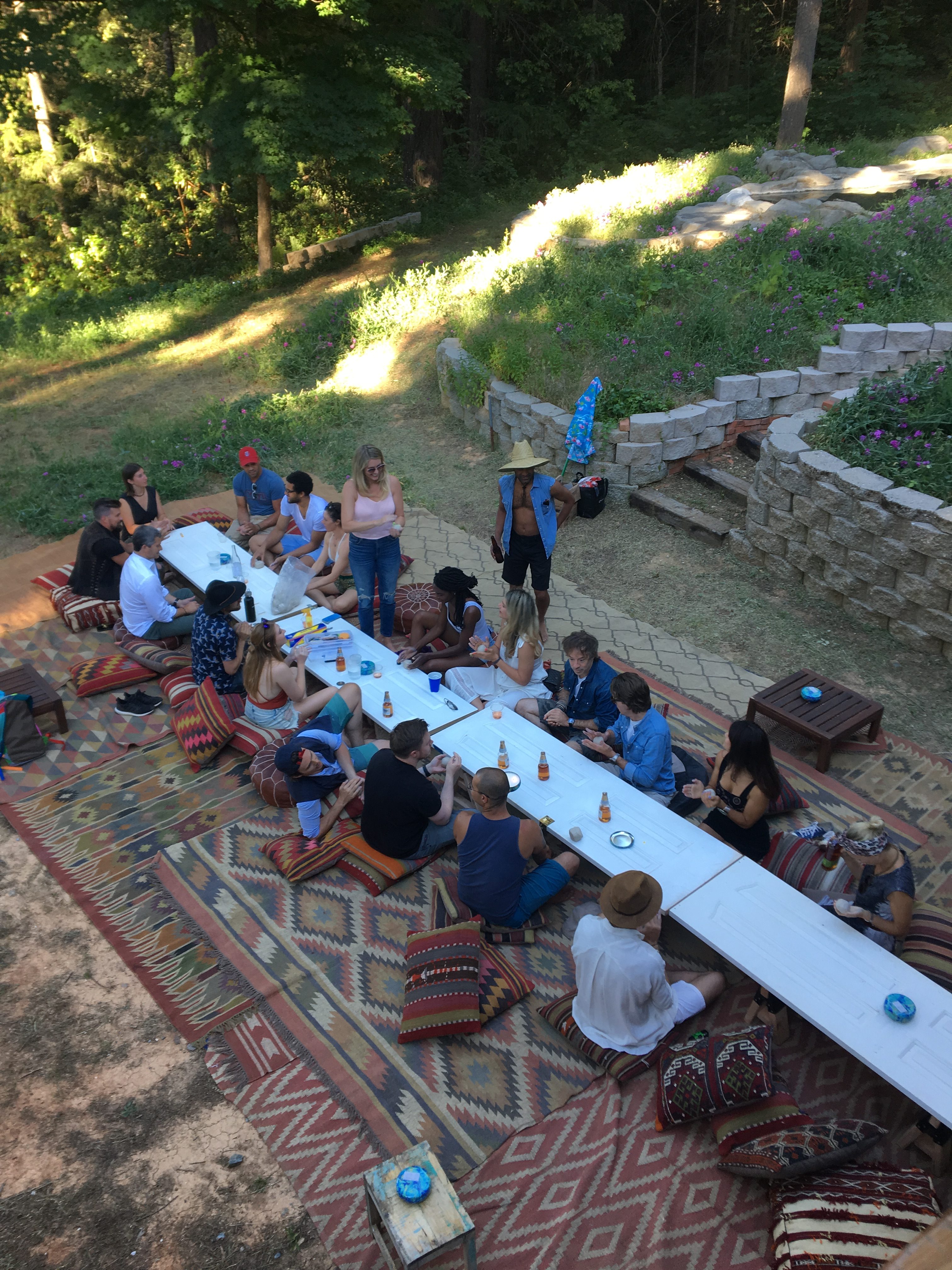 A group sits on cushions around a long, low table outdoors, enjoying food and drinks. They are surrounded by greenery and a stone retaining wall with vibrant flowers.