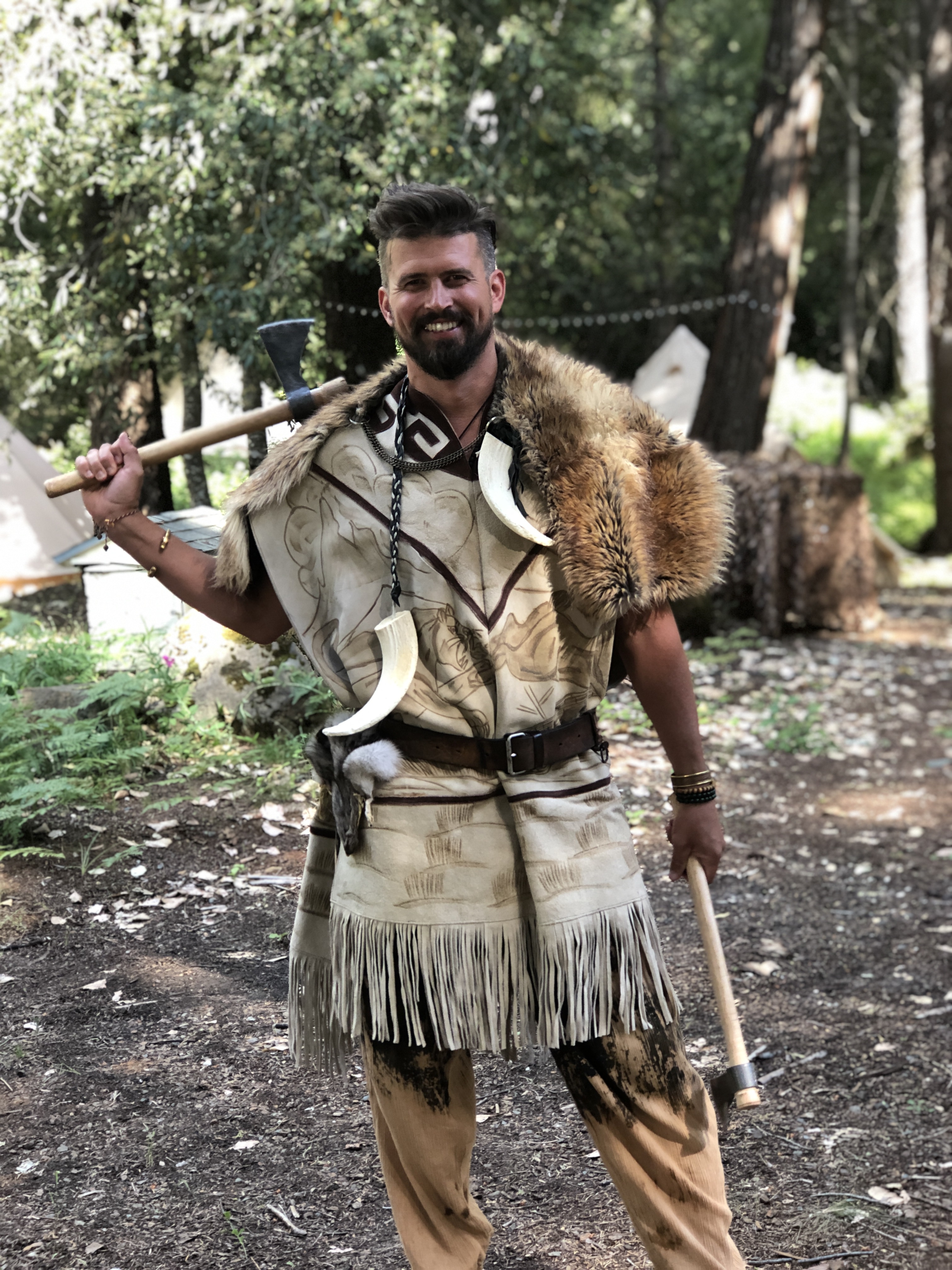 A man, smiling, stands outdoors in the forest wearing a fur cape, a fringed tunic, and animal tooth necklace, holding an axe and a mallet in each hand.