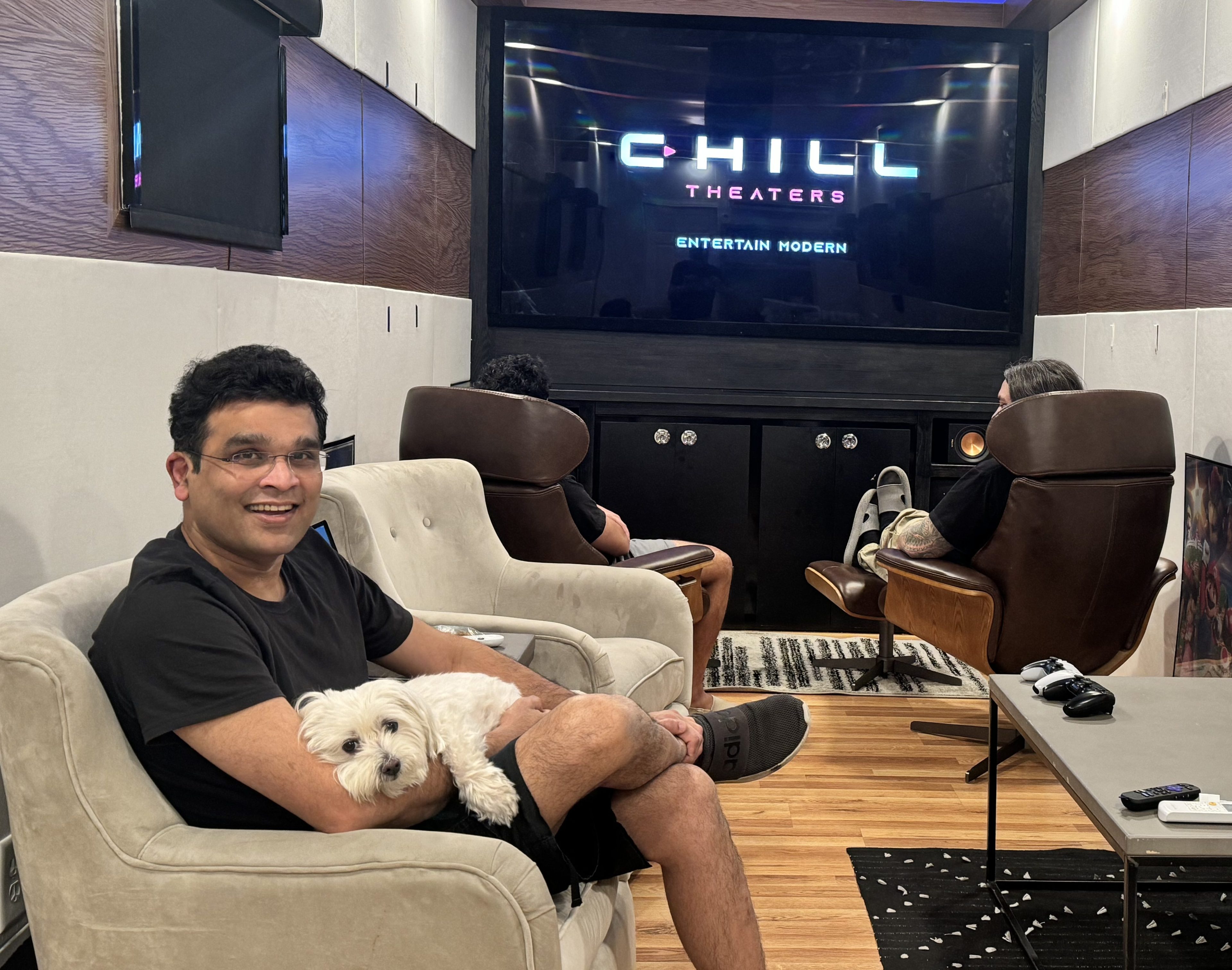 A man in glasses smiles, sitting on a beige chair with a small white dog. Two other people sit in brown chairs, watching a large screen displaying &quot;Chill Theaters.&quot;