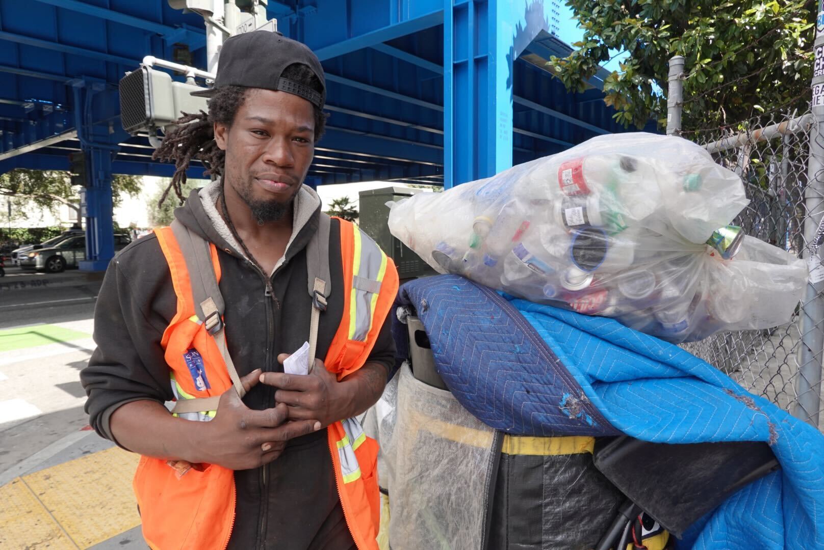 A man in an orange safety vest and black hoodie stands under a blue structure, holding papers. He is next to a cart loaded with a large bag of plastic bottles.