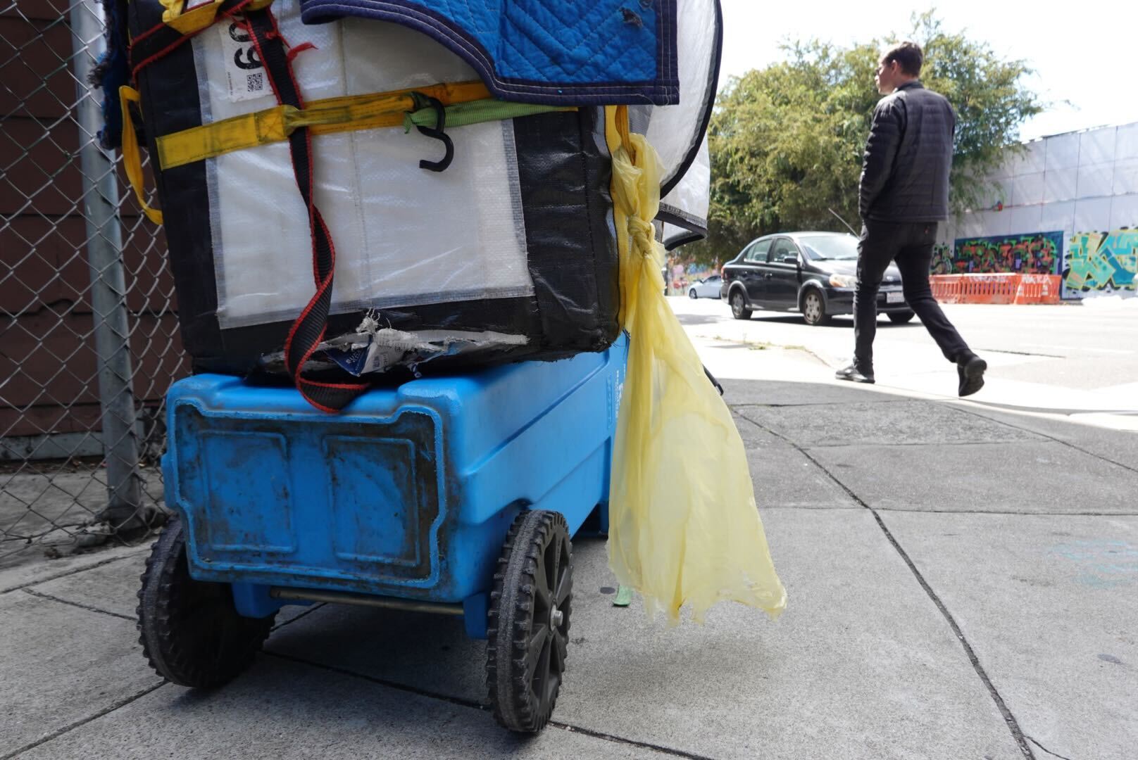 A loaded blue cart sits on a sidewalk next to a chain-link fence. A man in a black jacket is walking nearby, and a black car and trees are visible in the background.