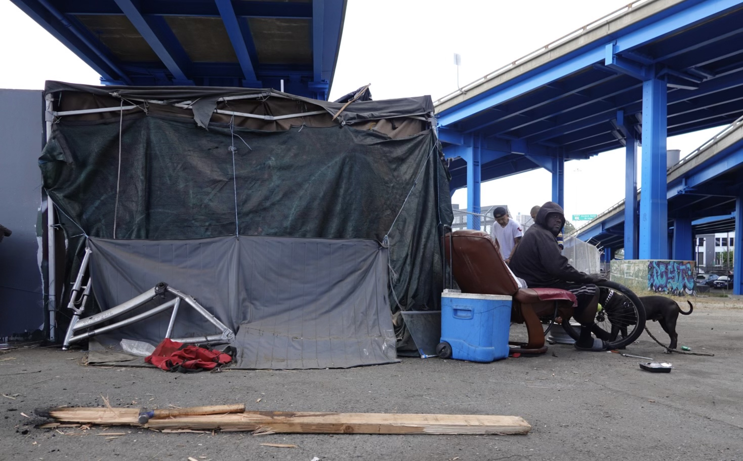 A man sits on a recliner under a blue overpass, next to makeshift shelter materials and a blue cooler. Another person and a dog are nearby on the concrete ground.