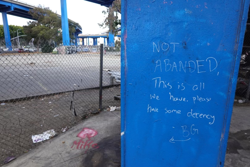 A blue structure has handwritten words, &quot;NOT ABANDED, This is all we have, please have some decency.&quot; Nearby, there is a fence, some graffiti, and trash.