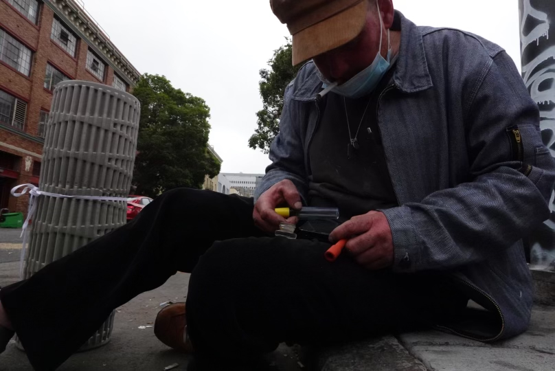 A man in a cap and denim jacket sits on a curb, looking down and holding tools. He is in an urban area with brick buildings and trees visible in the background.