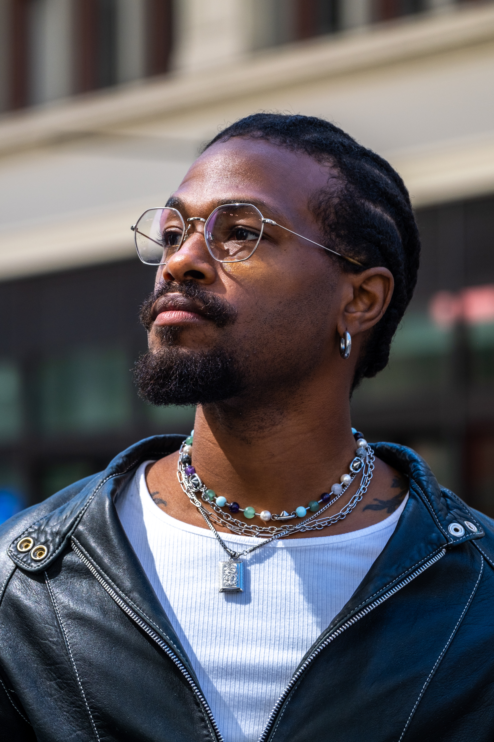 A man with glasses and braided hair is wearing a black leather jacket, a white shirt, and multiple necklaces. He has a contemplative expression and looks upwards.