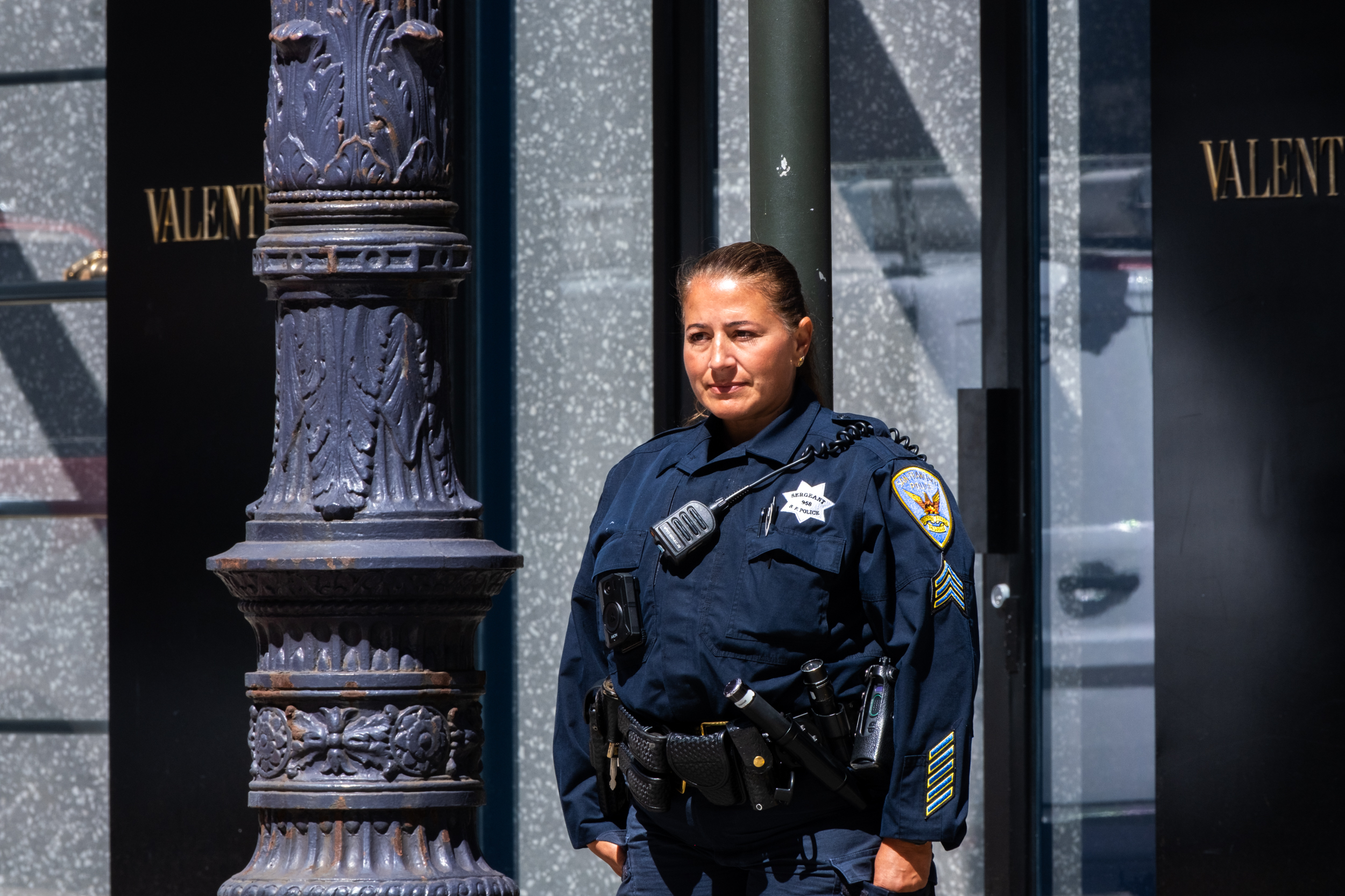 A female police officer in a dark blue uniform stands beside an ornate lamppost, with &quot;Valentino&quot; signage visible in the background.