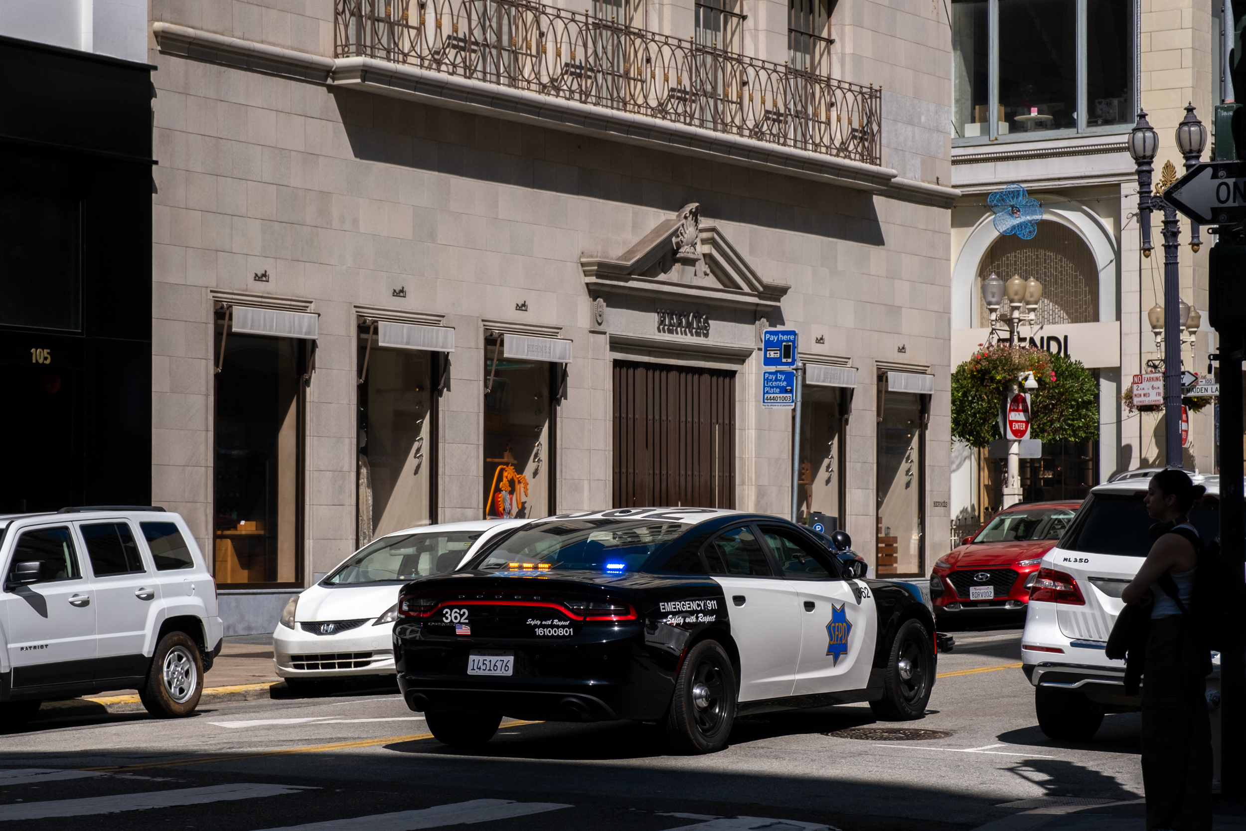 A police car with flashing lights is parked on a city street near high-end shops. Nearby are other parked vehicles, a pedestrian, and a parking meter sign.