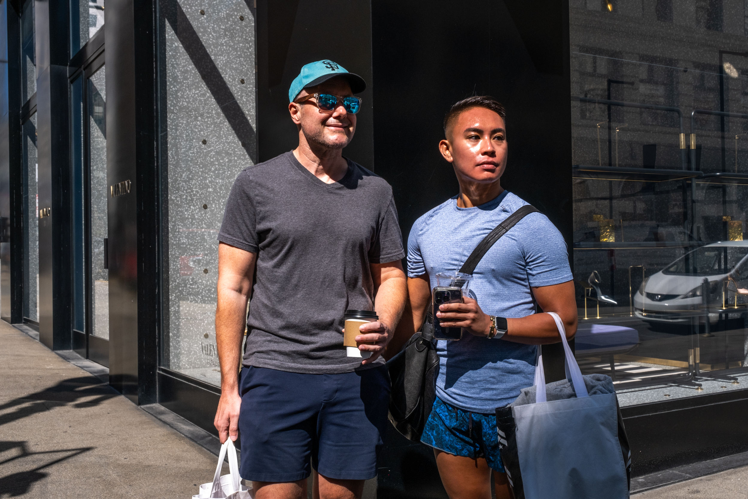 Two individuals stand outdoors, one in a blue hat and sunglasses holding a coffee cup, the other in a blue shirt holding an iced drink. They carry shopping bags.