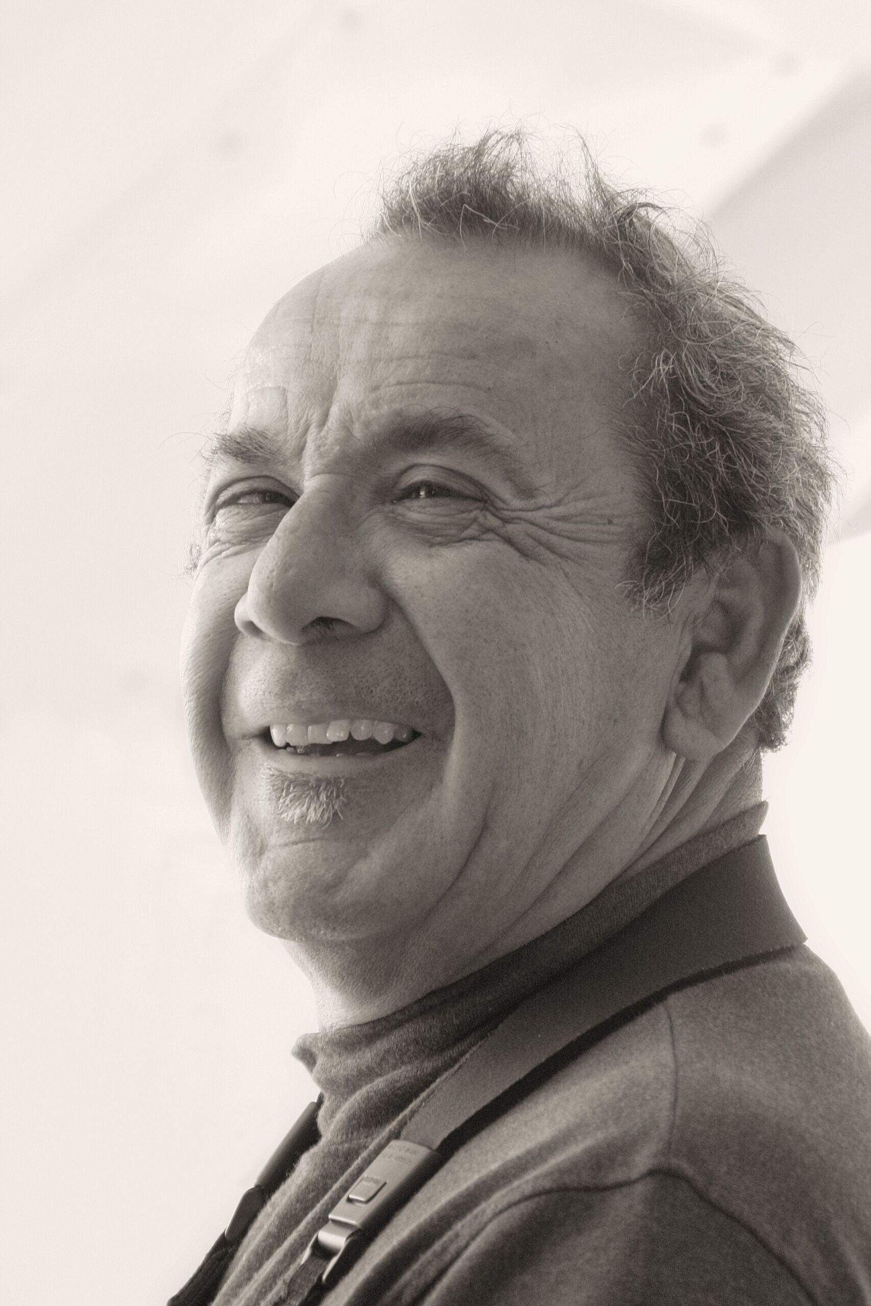 The image is a close-up black-and-white photo of an older man smiling warmly. He has short, wavy hair and is wearing a high-collared shirt with a strap around his neck.