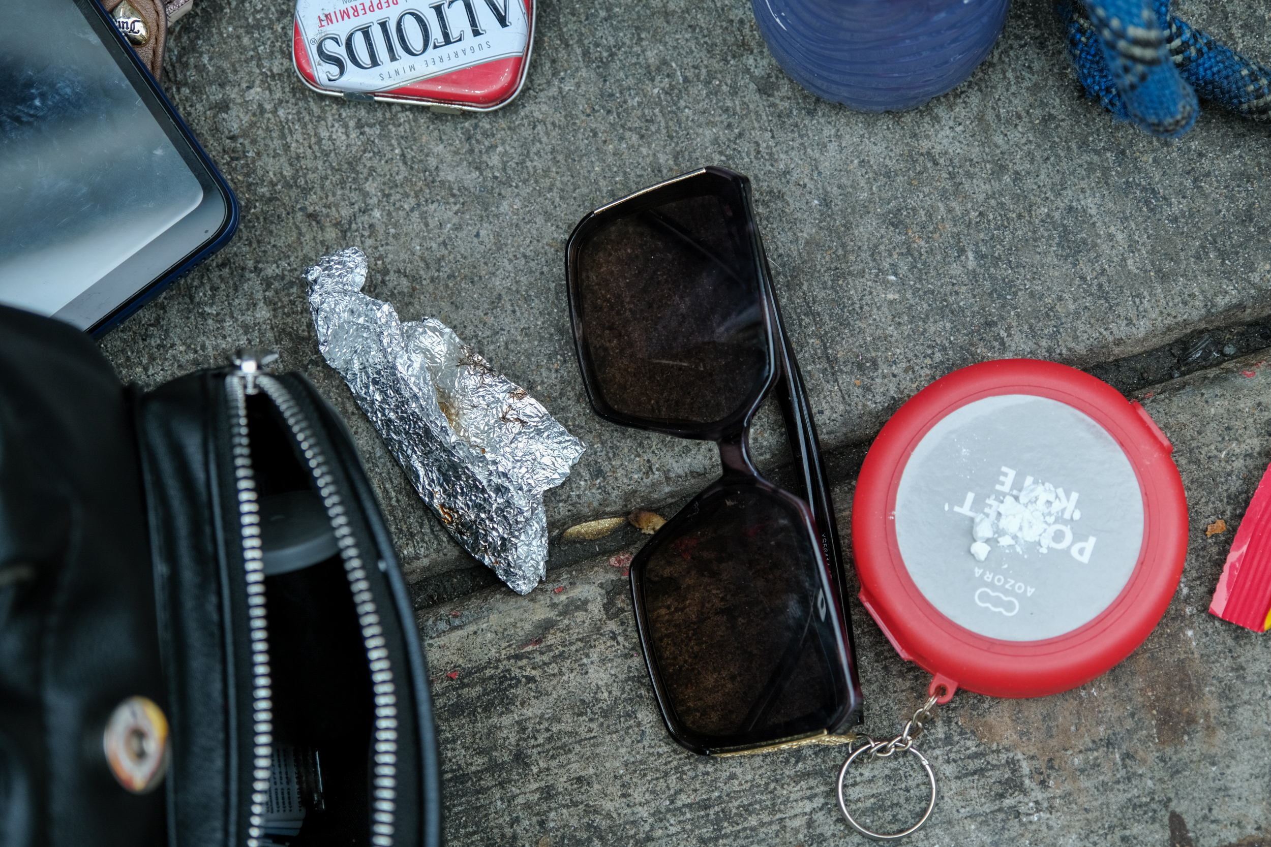 The image shows sunglasses, a crumpled piece of foil, an opened tin of Altoids, a small flat circular container, and part of a black bag on a concrete surface.