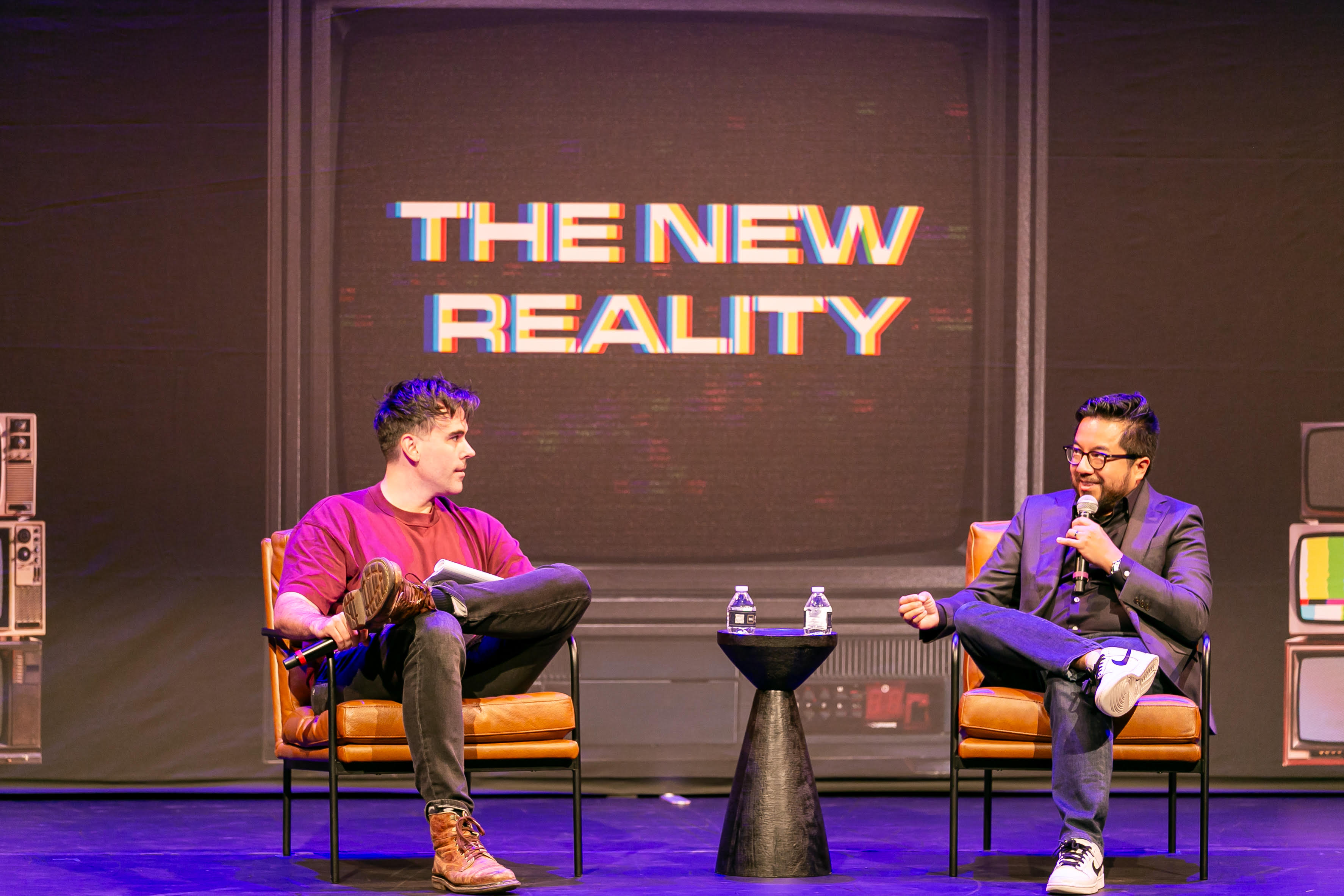 Two men sit on stage in leather chairs, talking. Behind them is a large screen displaying "THE NEW REALITY." They're engaged in conversation, with microphones and water bottles on a small table between them.