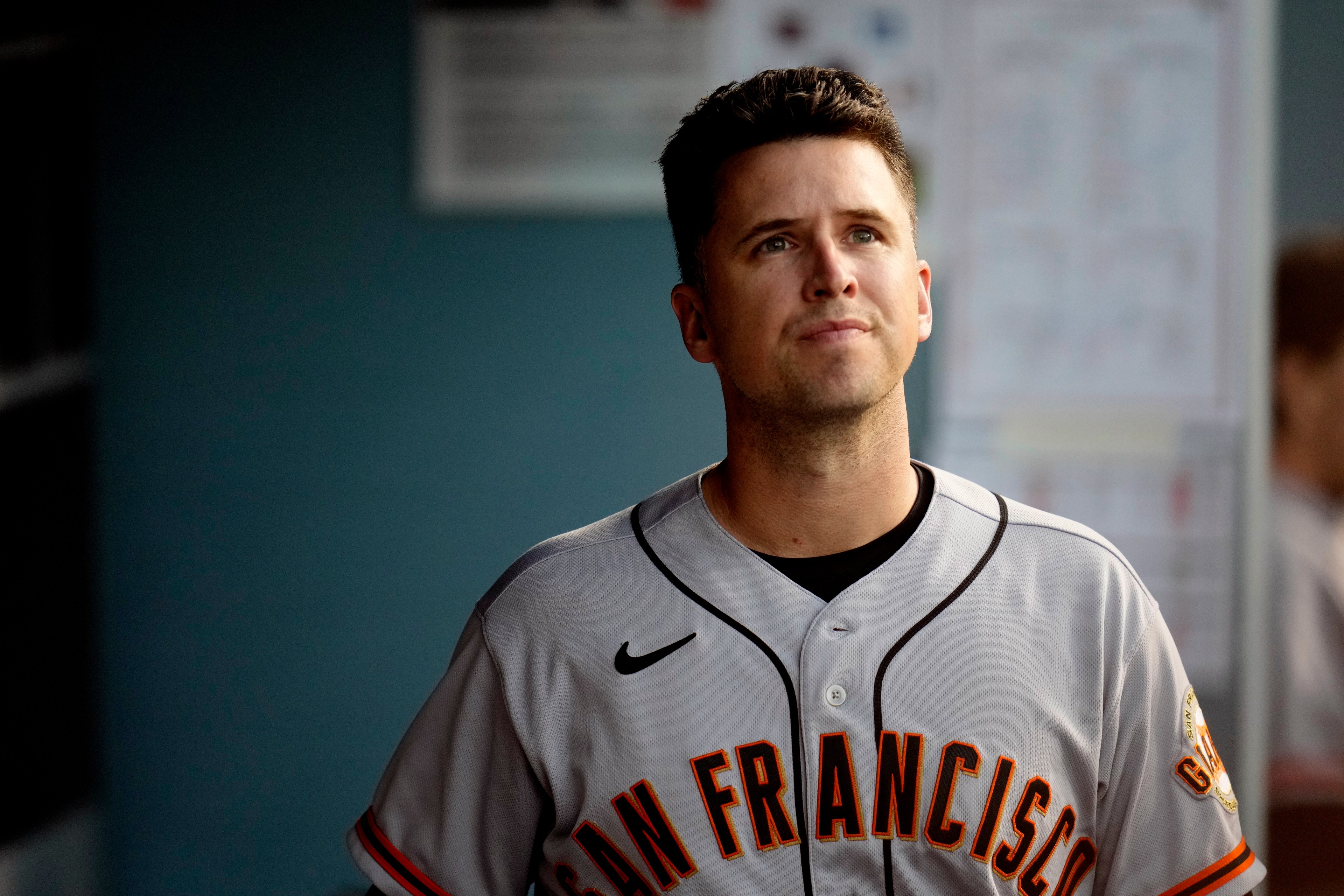 A baseball player in a San Francisco jersey looks upwards while standing in a dugout, with blurred background elements and natural lighting.