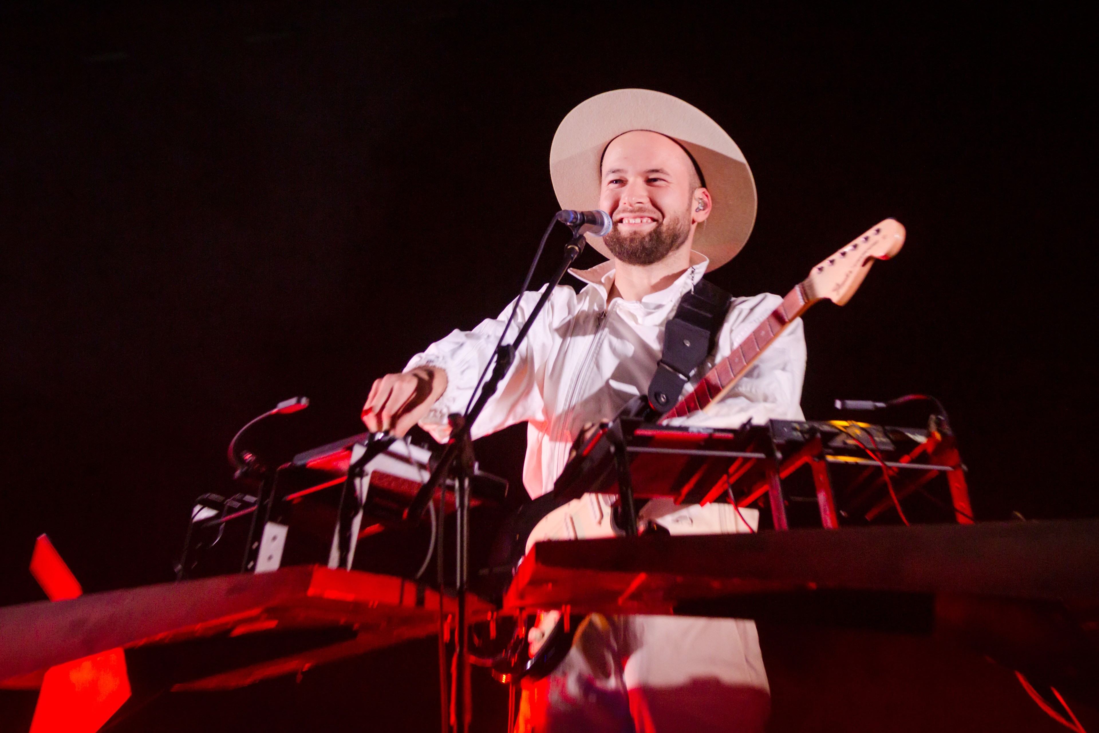 A man behind a microphone is smiling while playing an electronic keyboard setup and a guitar on stage, wearing a wide-brimmed hat and a white outfit.