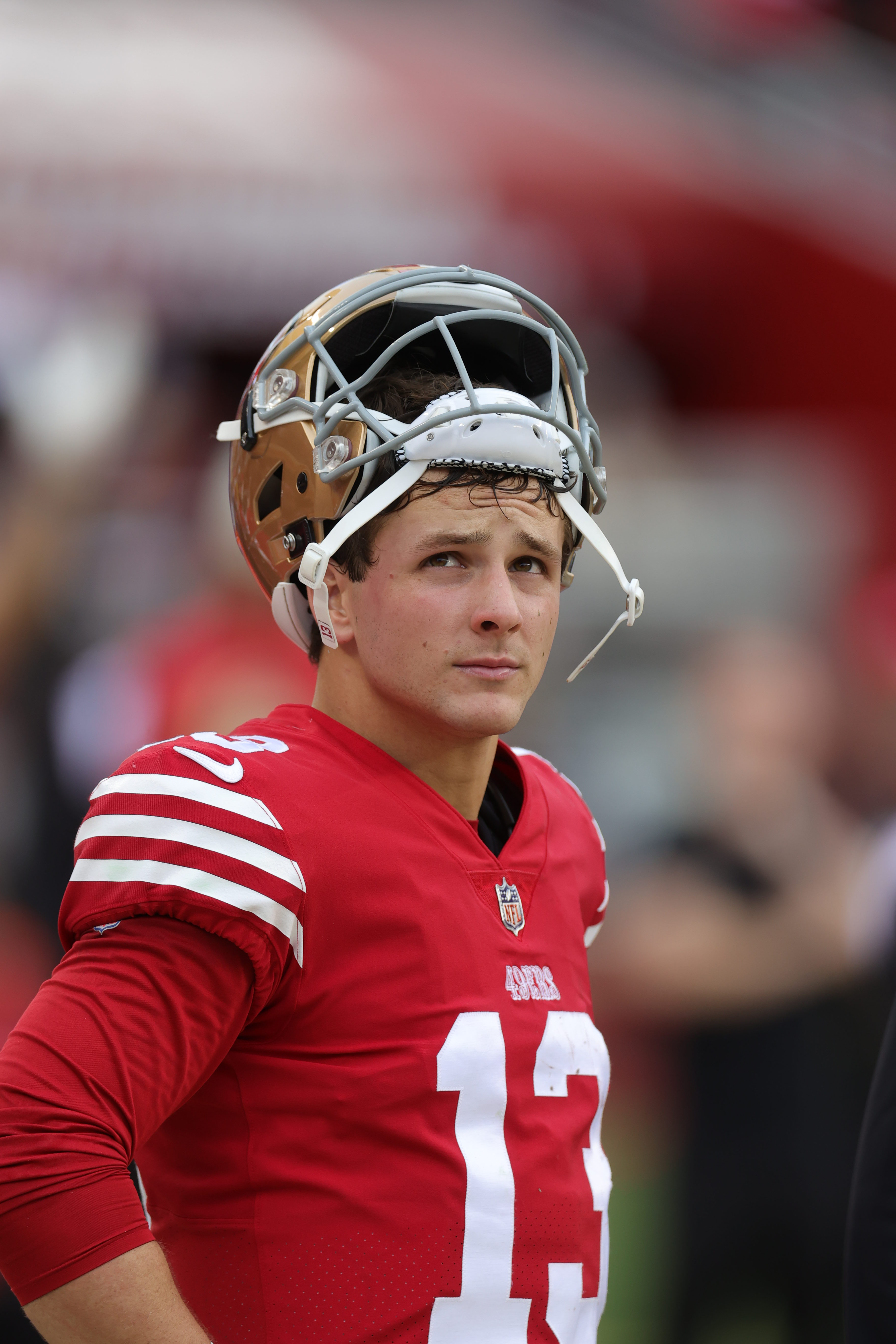 A football player wearing a red and white uniform stands with his helmet resting on top of his head, looking slightly upward and to the side.