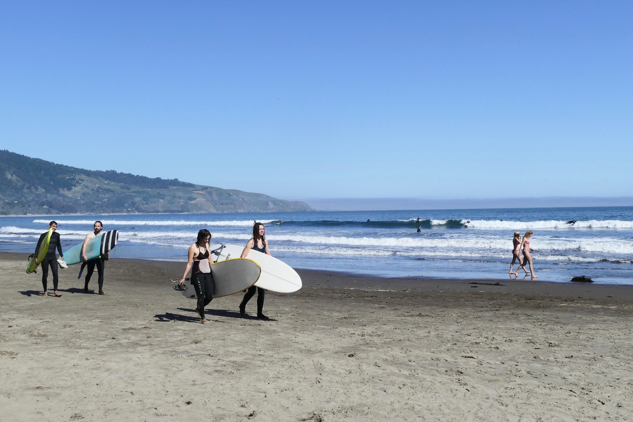 Surfers walk along the beach carrying white surfboards. In the background are mountains.