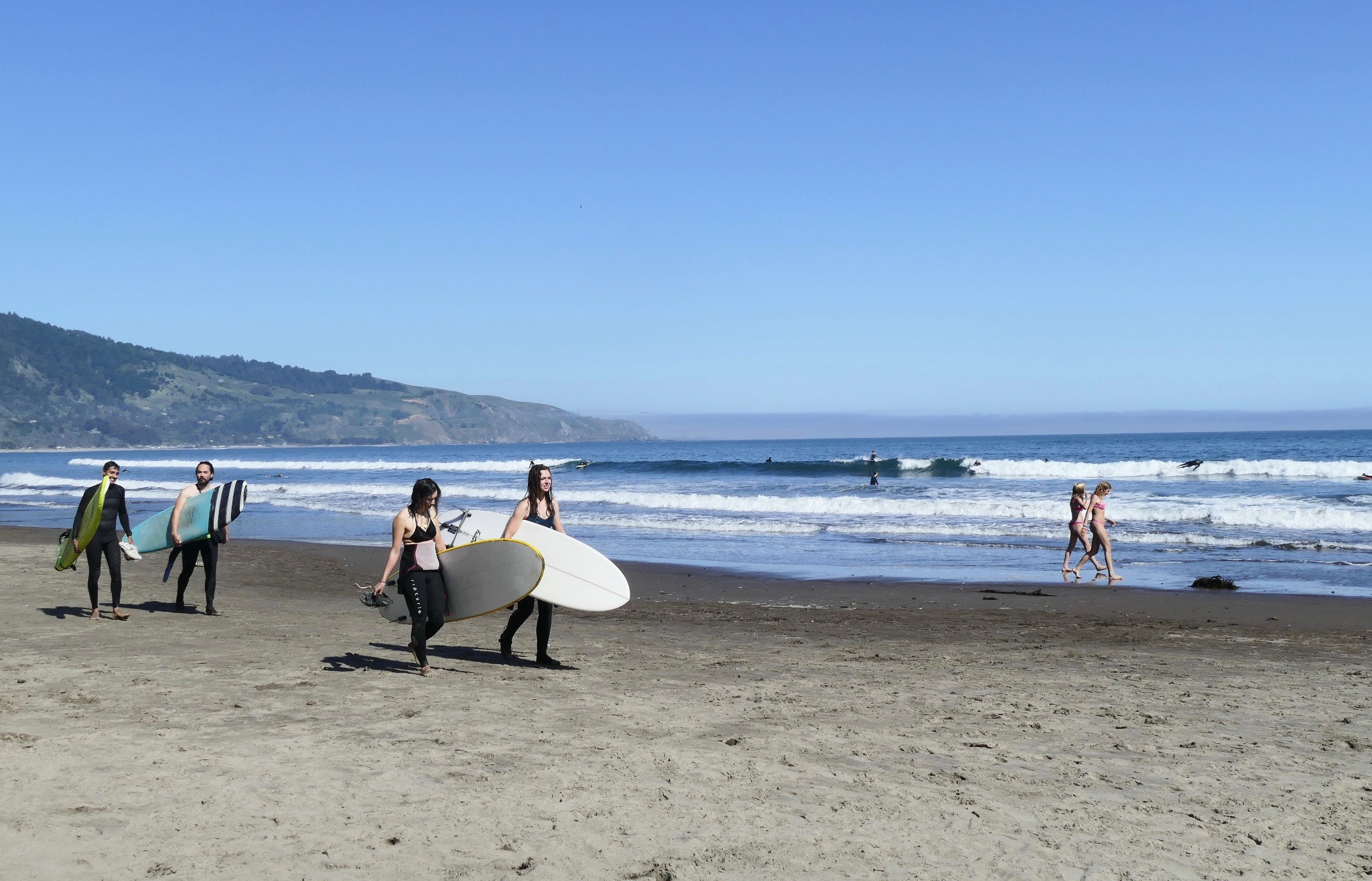 Surfers walk along the beach carrying white surfboards. In the background are mountains.