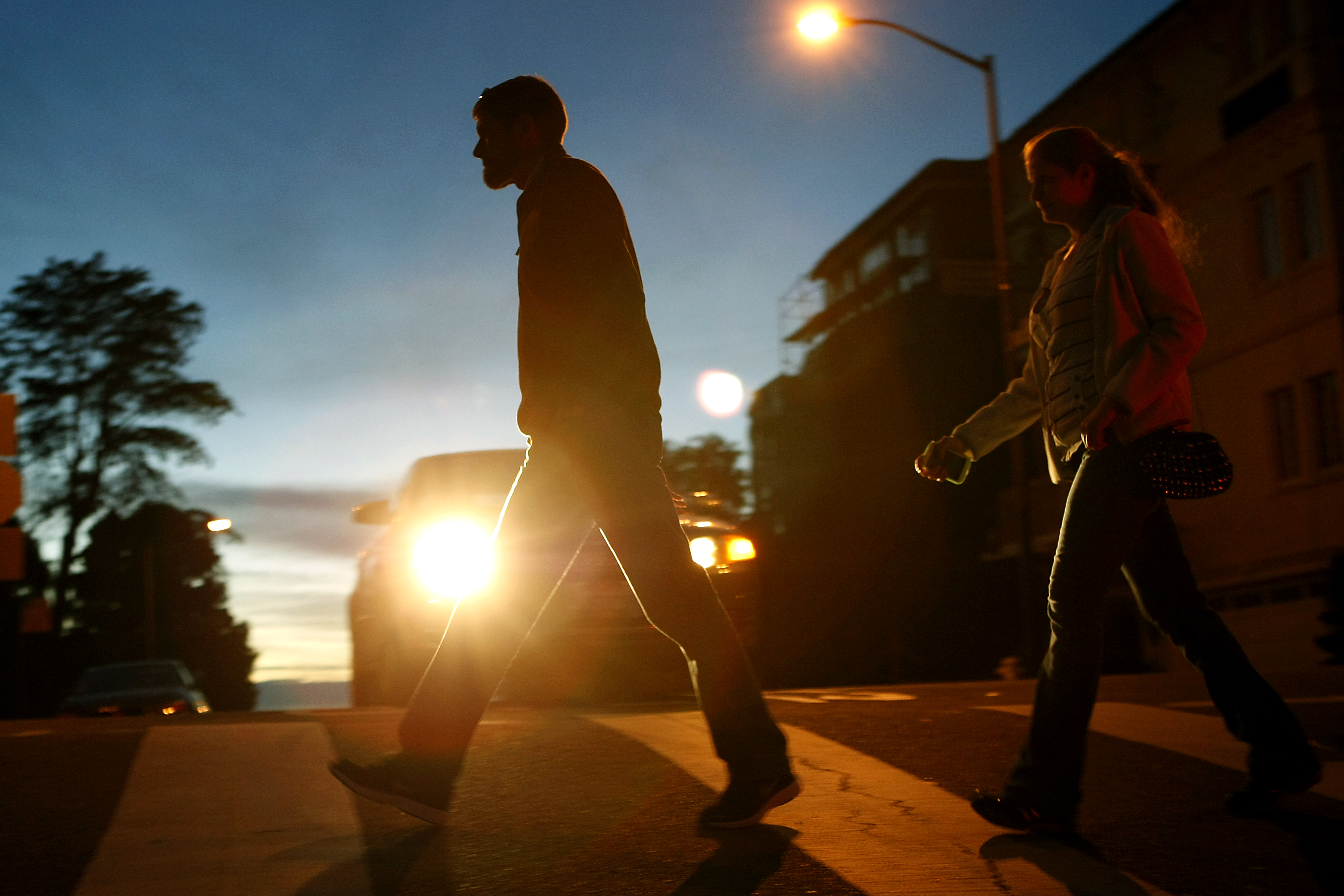 Two people are walking across a street at dusk. A car's headlights shine brightly behind them, silhouetting their figures against the evening sky.