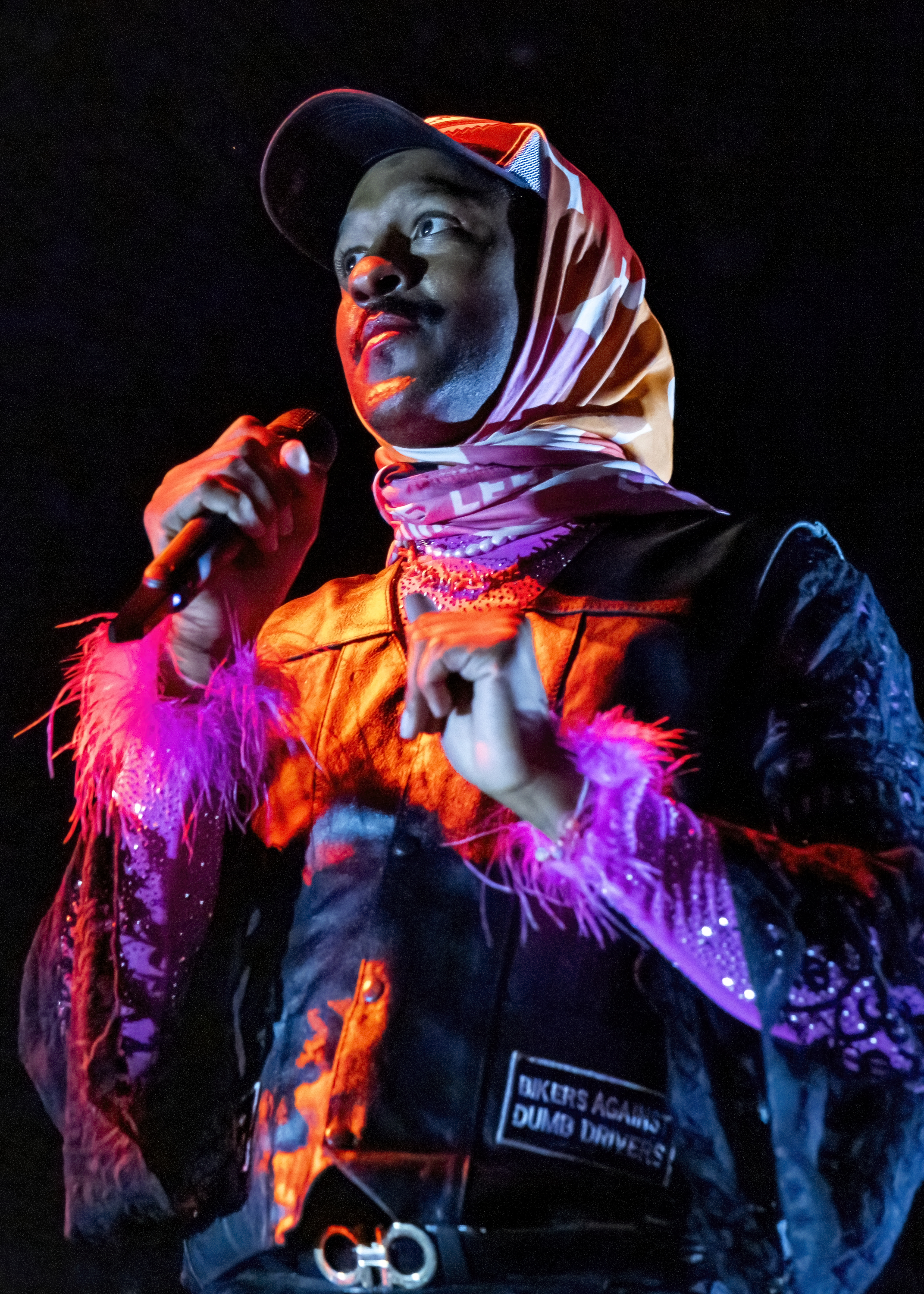 A person on stage holds a microphone, wearing a baseball cap, a patterned headscarf, a sparkly jacket with pink fluffy trim, and a patch that reads &quot;BIKERS AGAINST DUMB DRIVERS.&quot;