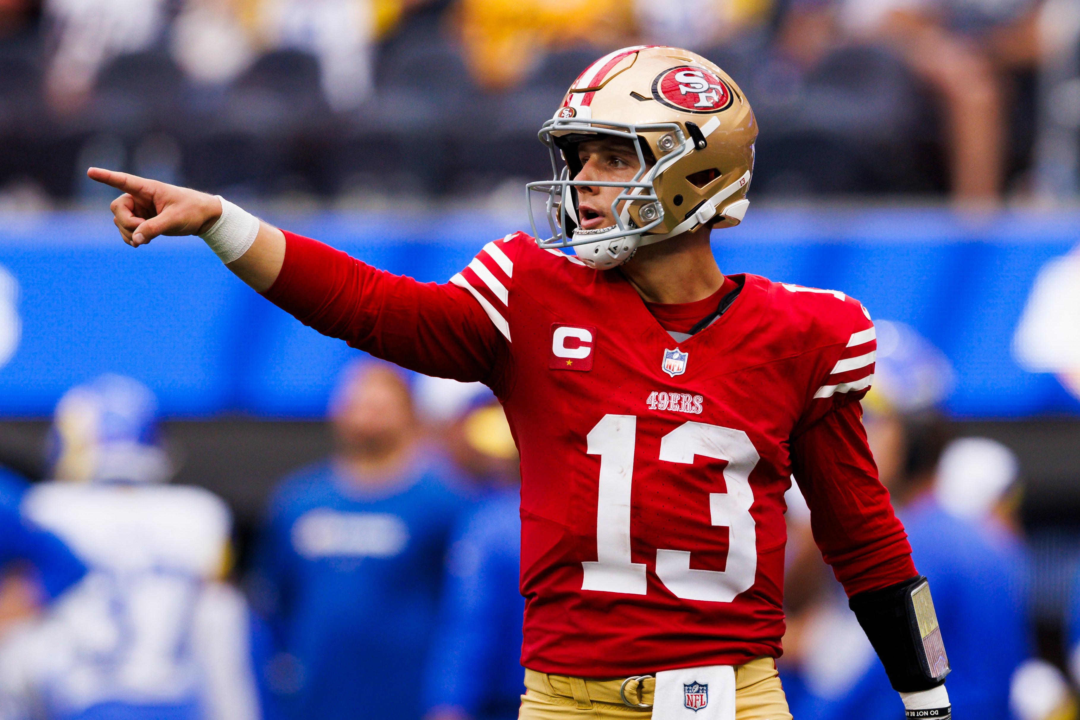 A football player in a red jersey and gold pants is on the field, pointing with his right arm extended. He wears a helmet and wristbands with a white towel tucked in.