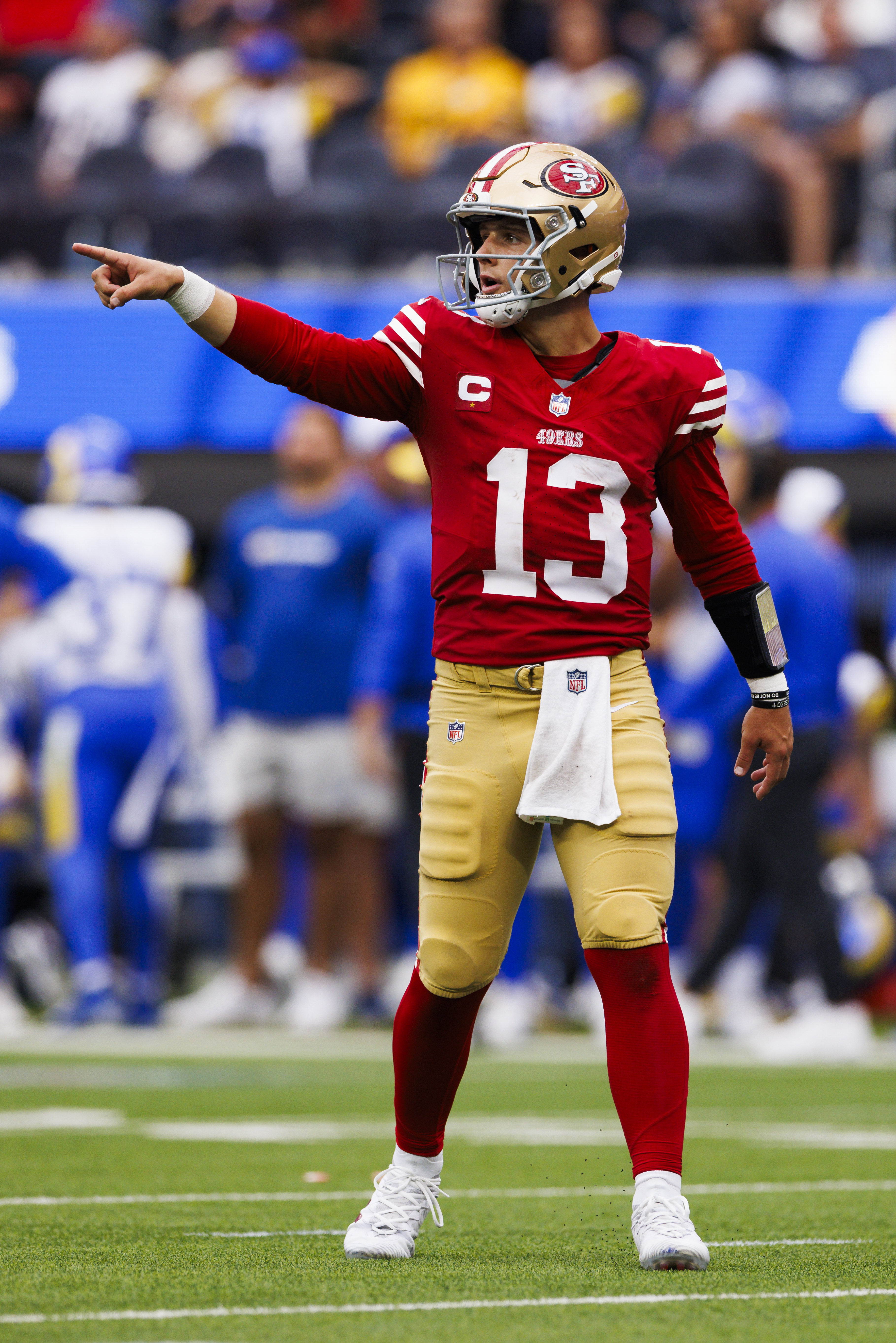 A football player in a red jersey and gold pants is on the field, pointing with his right arm extended. He wears a helmet and wristbands with a white towel tucked in.