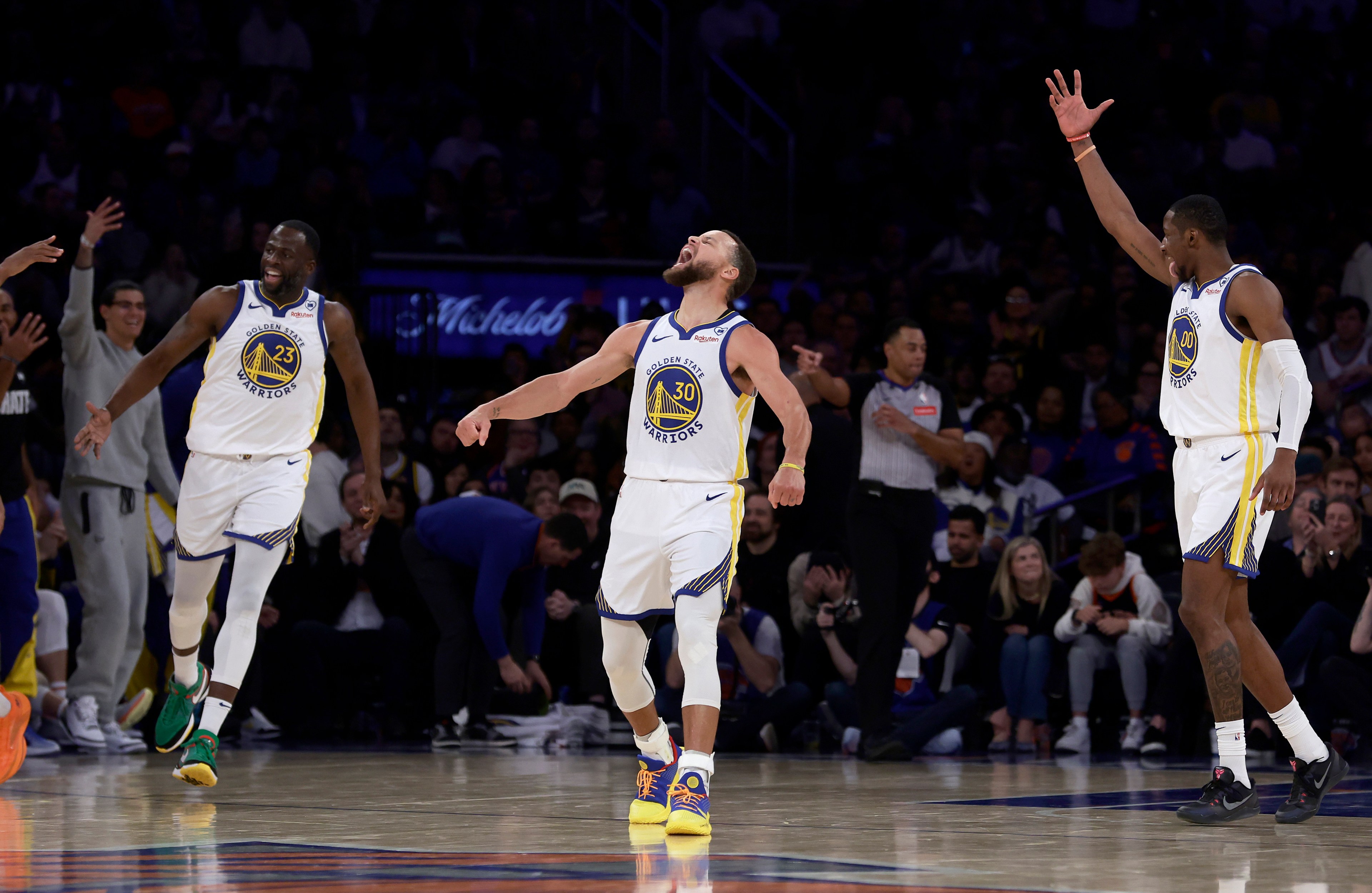 Three basketball players, wearing Golden State Warriors jerseys, celebrate energetically on the court as an official stands nearby and a crowd watches in the background.