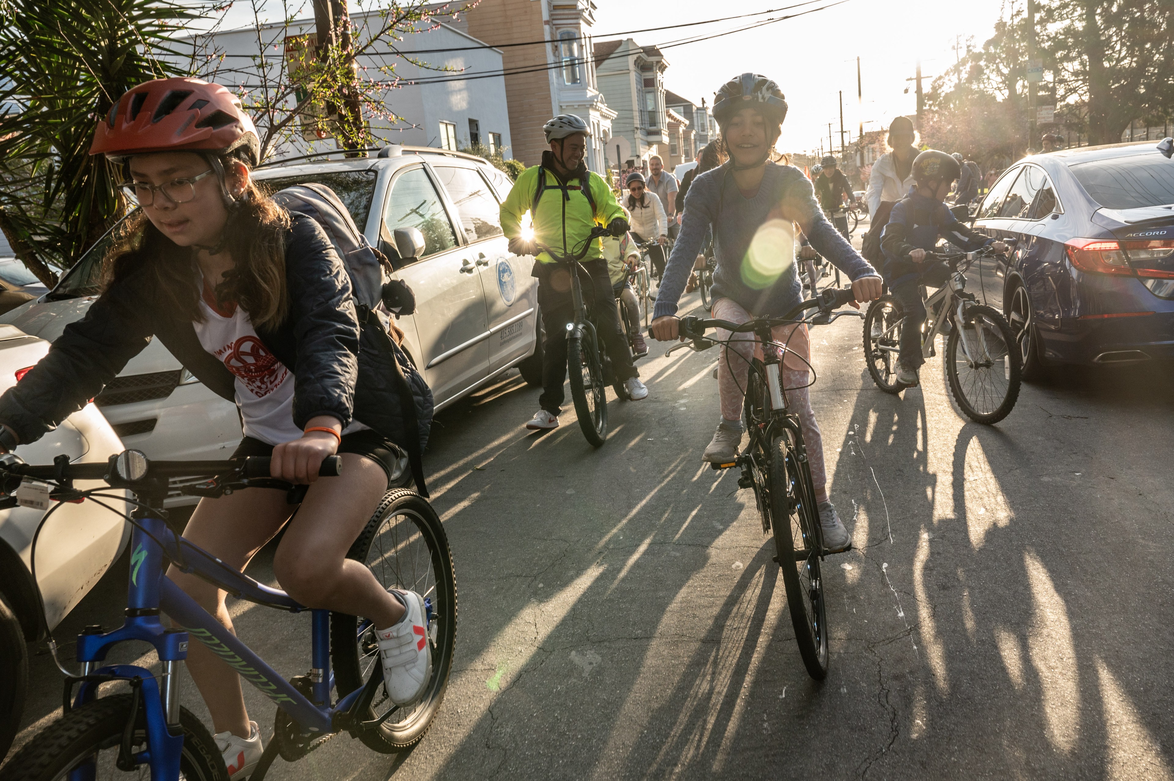 A group of people of various ages is riding bicycles on a street, surrounded by parked cars on either side, under the setting sun casting long shadows.