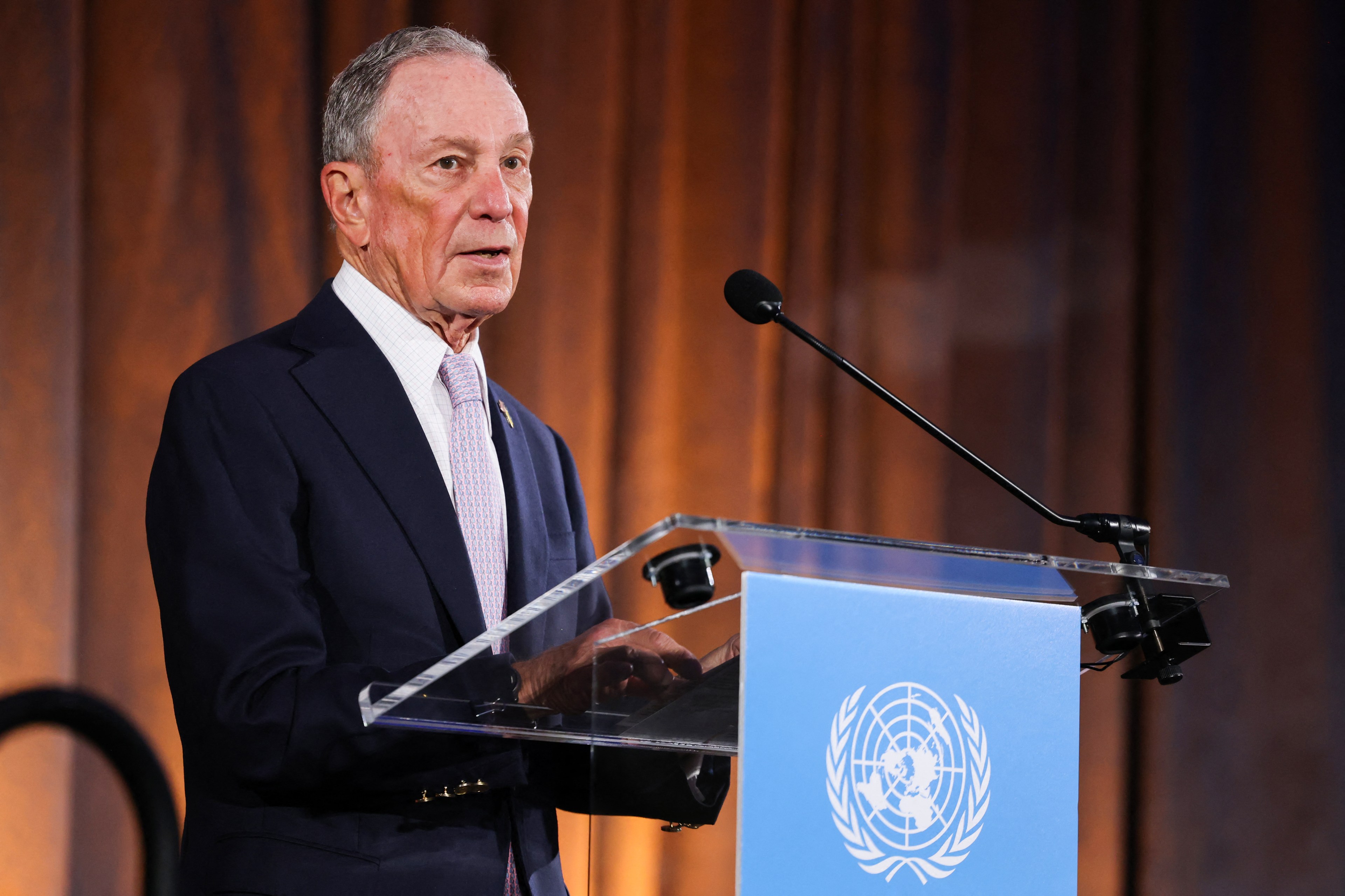 An older man in a suit and tie is speaking at a podium with a United Nations emblem. The background is a brownish curtain.