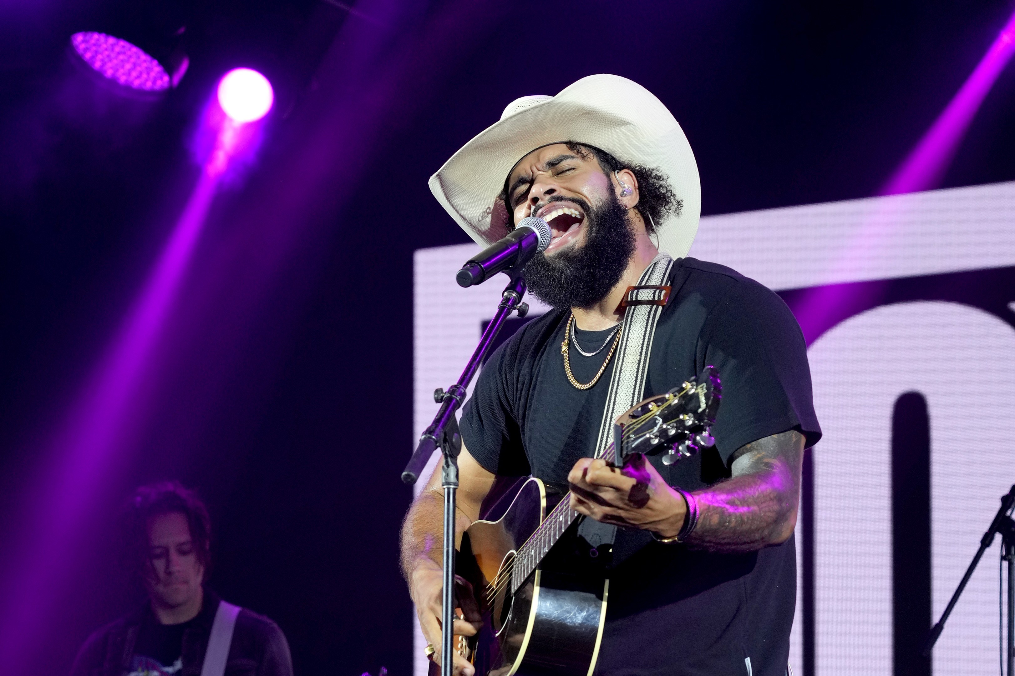 A bearded man in a cowboy hat passionately sings and plays acoustic guitar on stage under purple lights, with a bassist playing in the background.