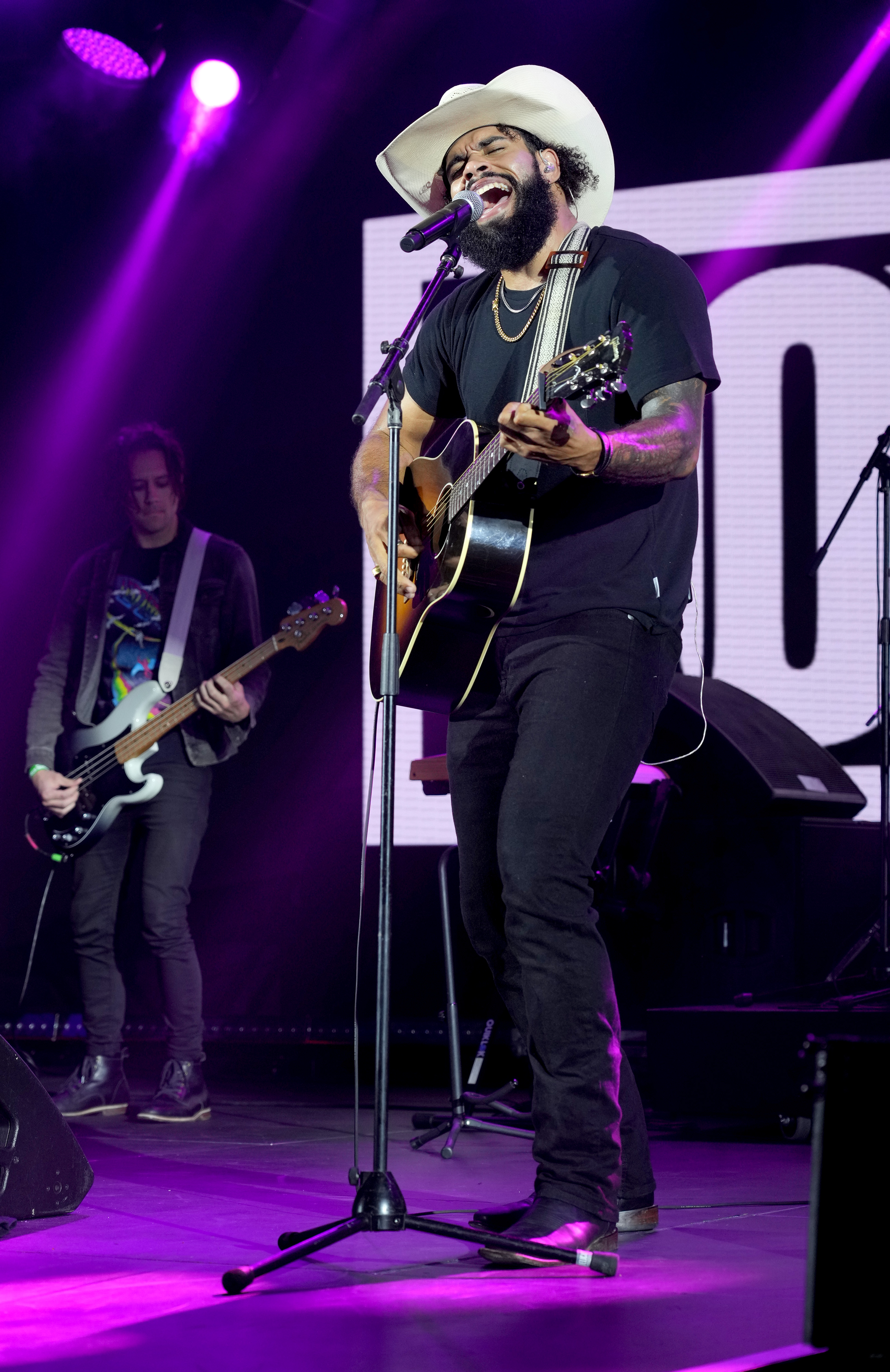 A bearded man in a cowboy hat passionately sings and plays acoustic guitar on stage under purple lights, with a bassist playing in the background.