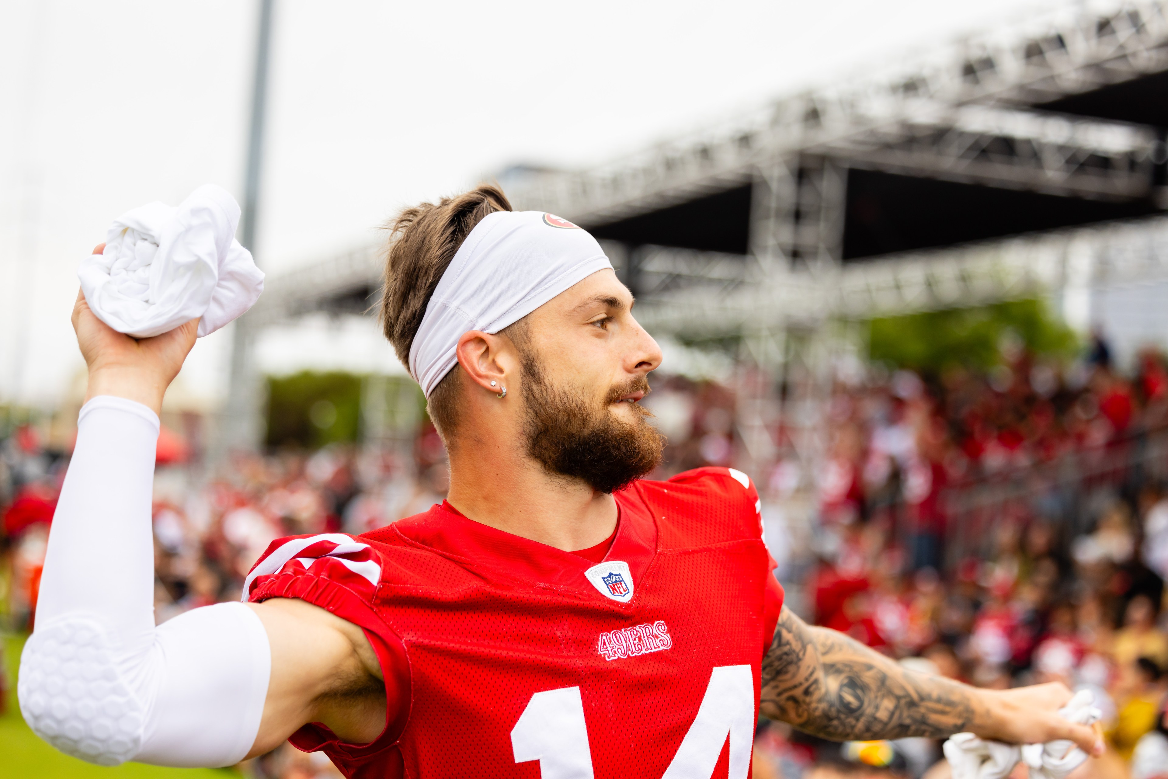 A football player in a red jersey, wearing a white headband and arm sleeve, throws a towel toward the audience at what appears to be a training event or game.