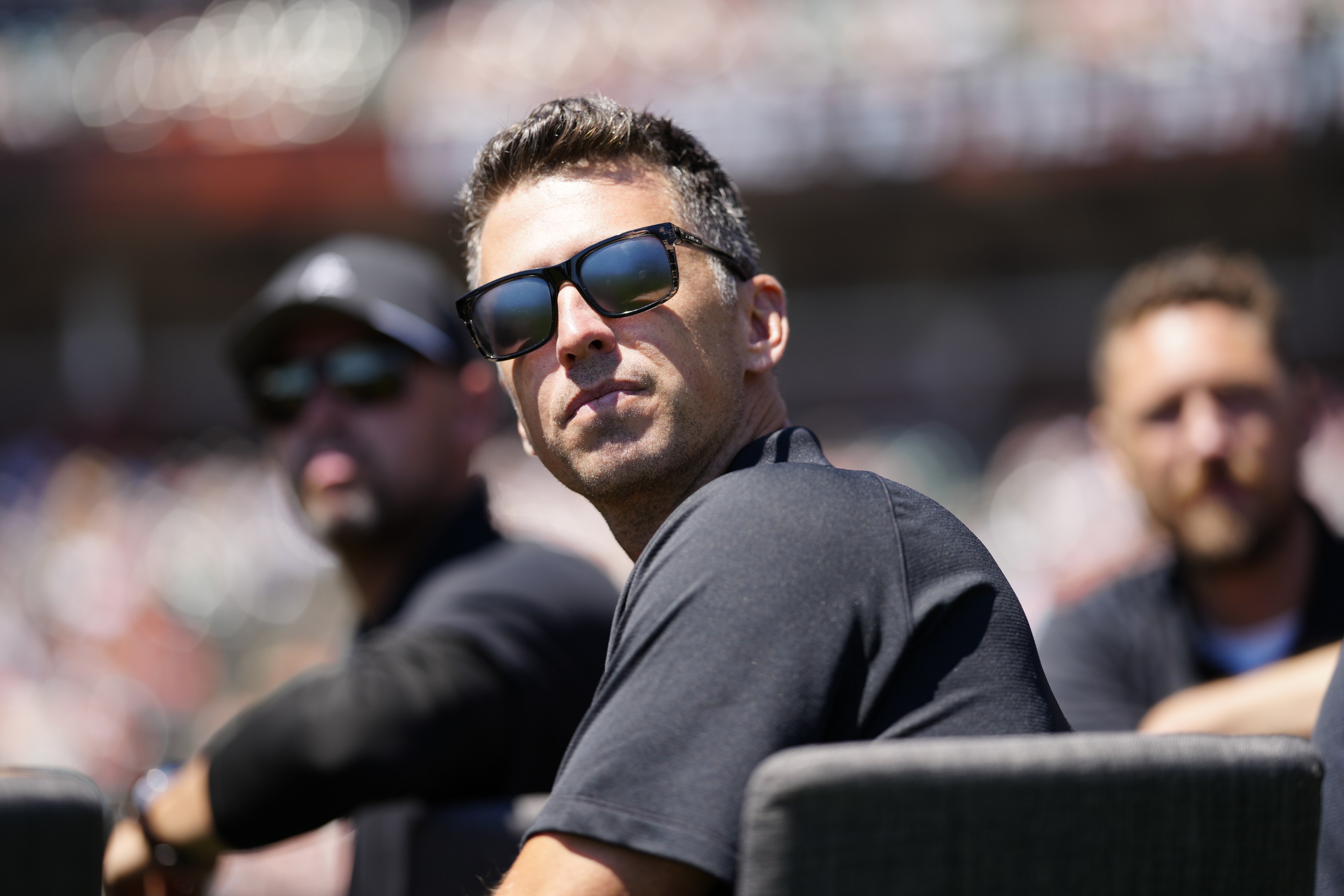 Three men wearing sunglasses and dark shirts are seated outdoors, with the central figure looking directly at the camera while the other two are slightly out of focus.