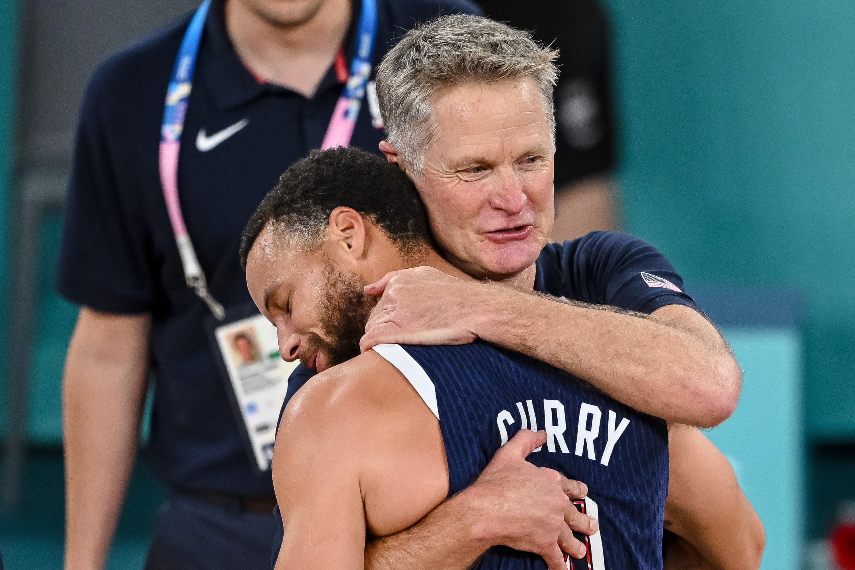 A man wearing a jersey labeled "CURRY" is being hugged by an older man in a coaching outfit. Another man stands in the background, watching them.