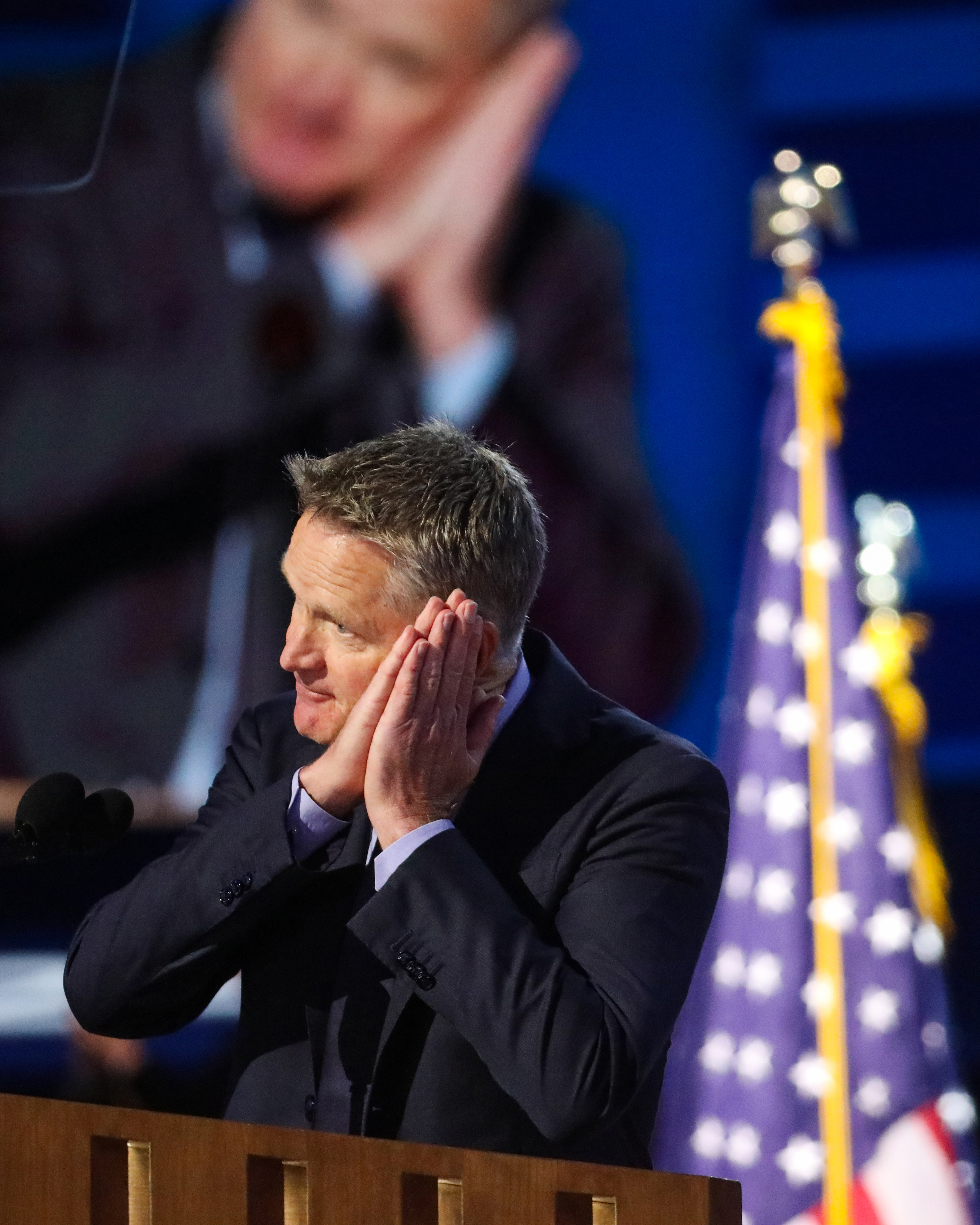 A man in a suit stands at a podium, hands pressed to his cheek in a sleeping gesture, with an American flag beside him. His large projected image appears behind.