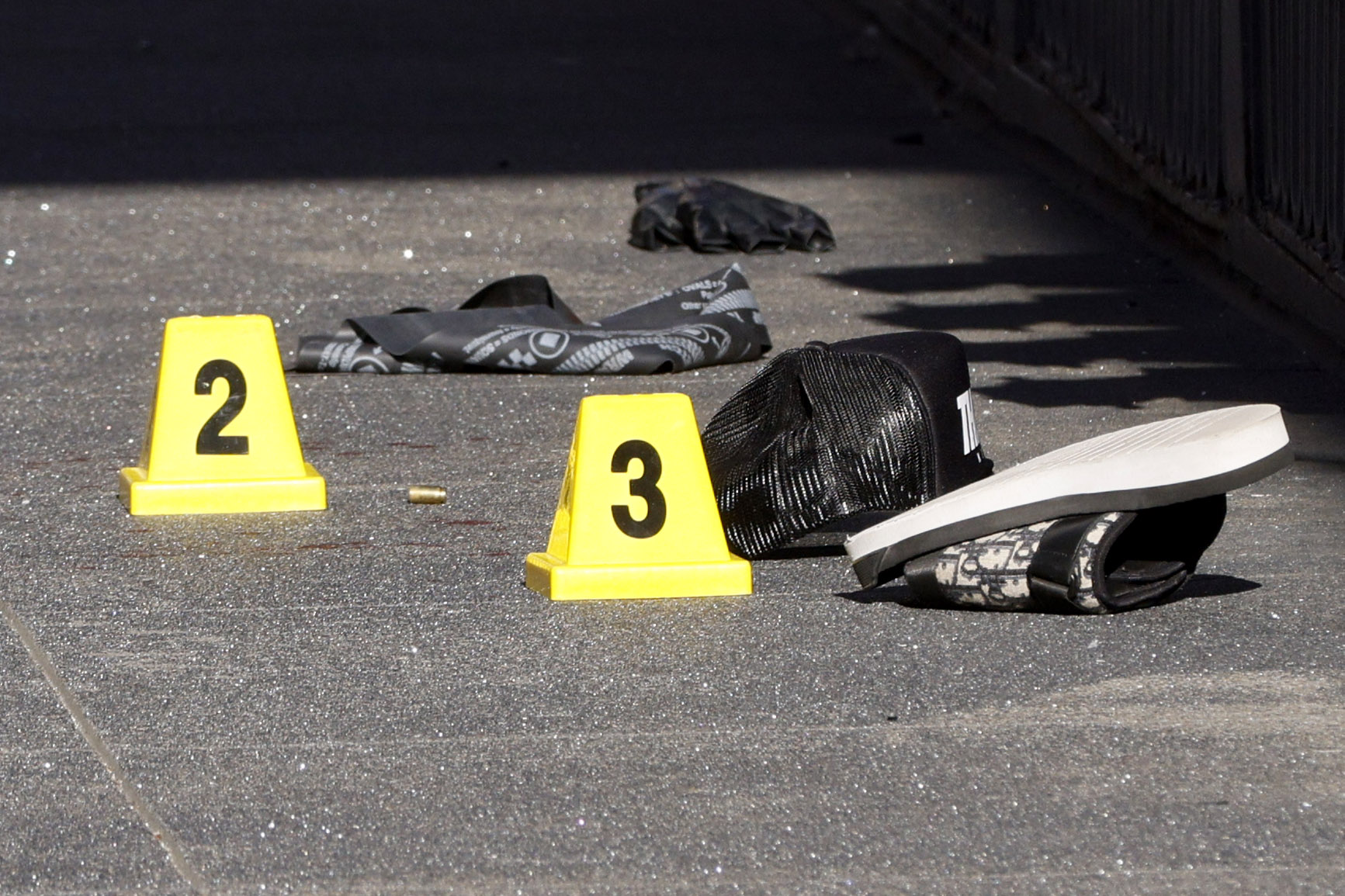 On a paved surface, there are yellow evidence markers with numbers 2 and 3. Nearby are a sandal, a cap, a cloth, and a bullet.