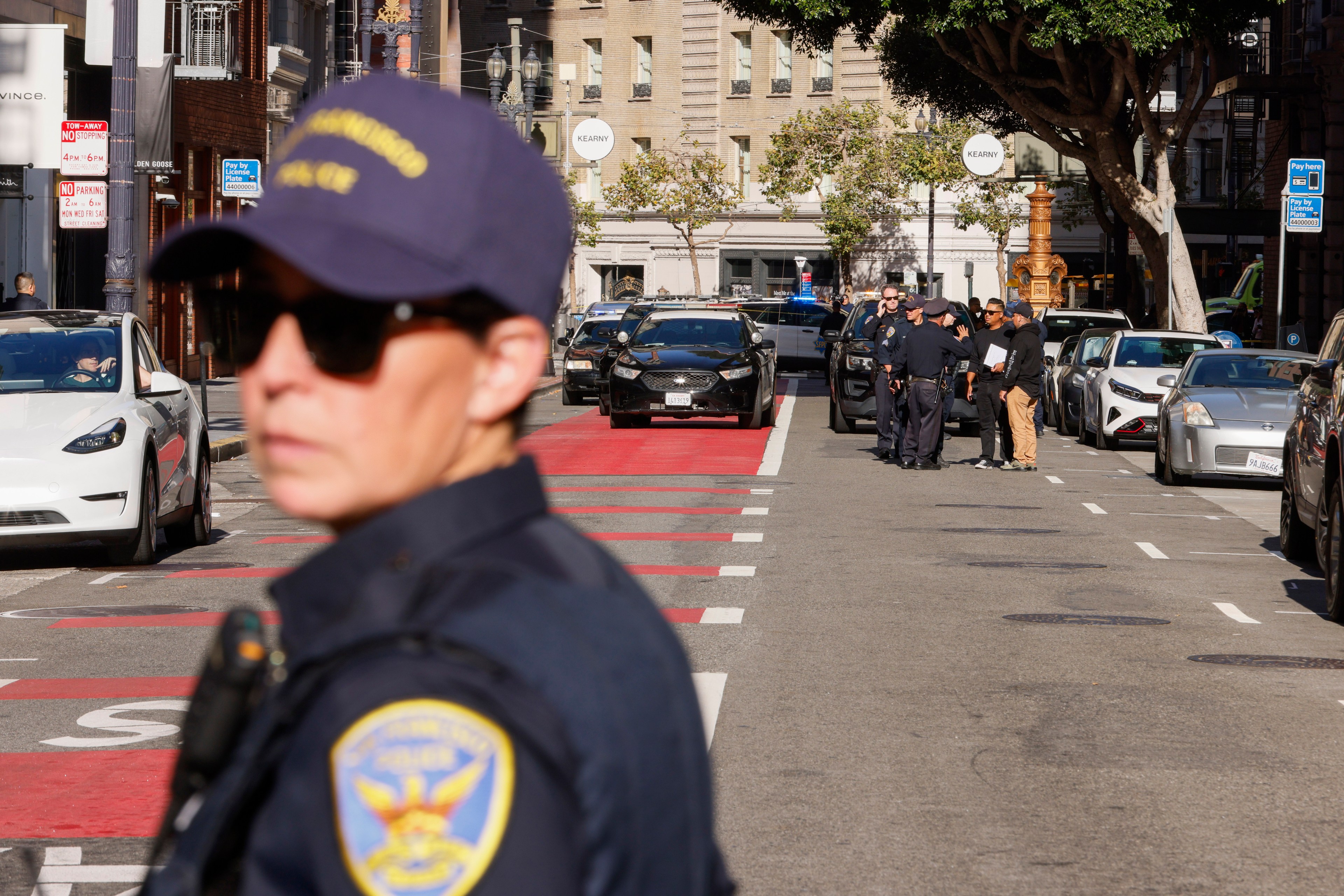 A police officer stands in focus in the foreground while other officers gather around individuals near parked cars in the background on a city street.