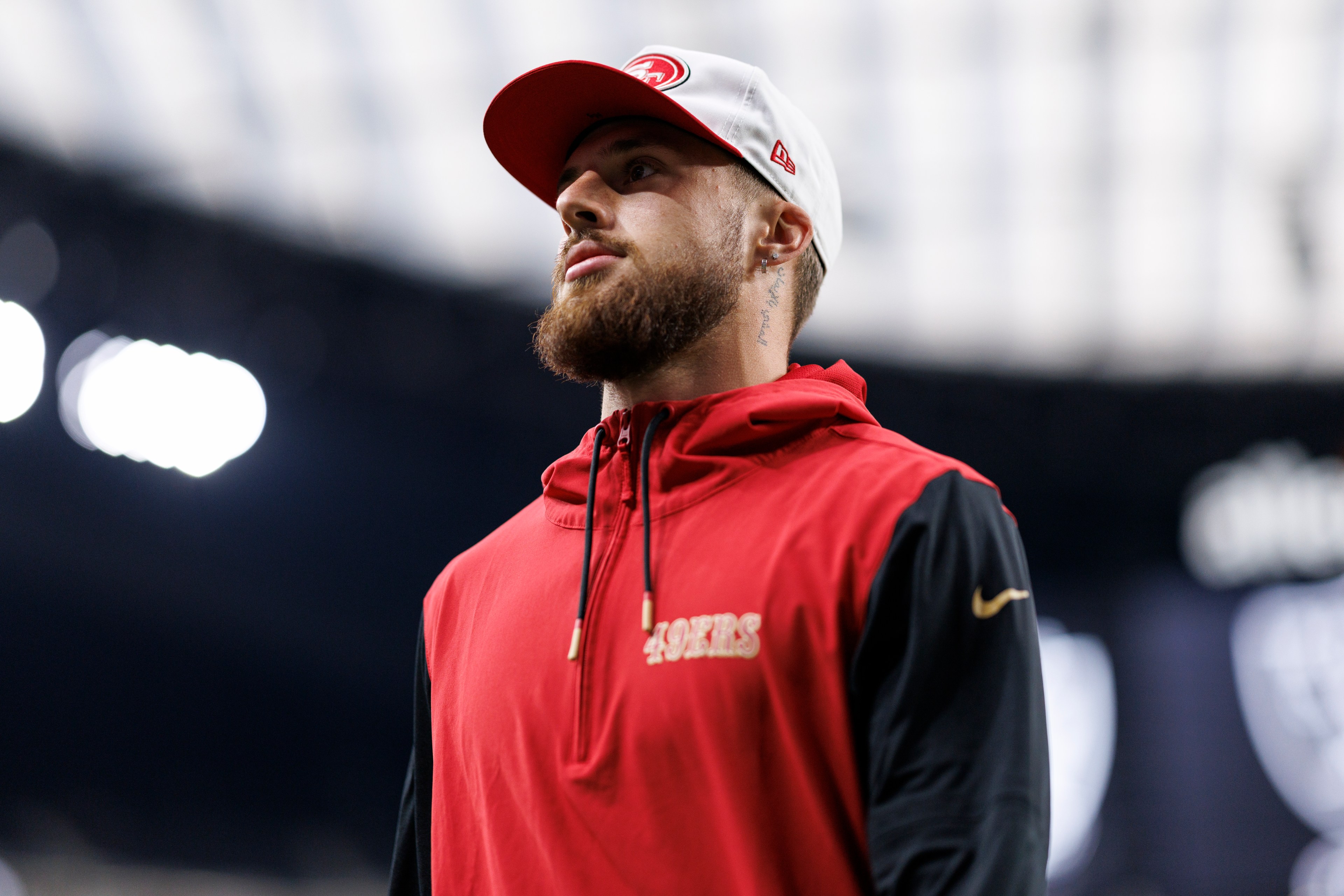 A bearded man wearing a white cap and a red sports jacket with "49ERS" on it stands in a stadium, looking up. The background is blurred with bright overhead lights.