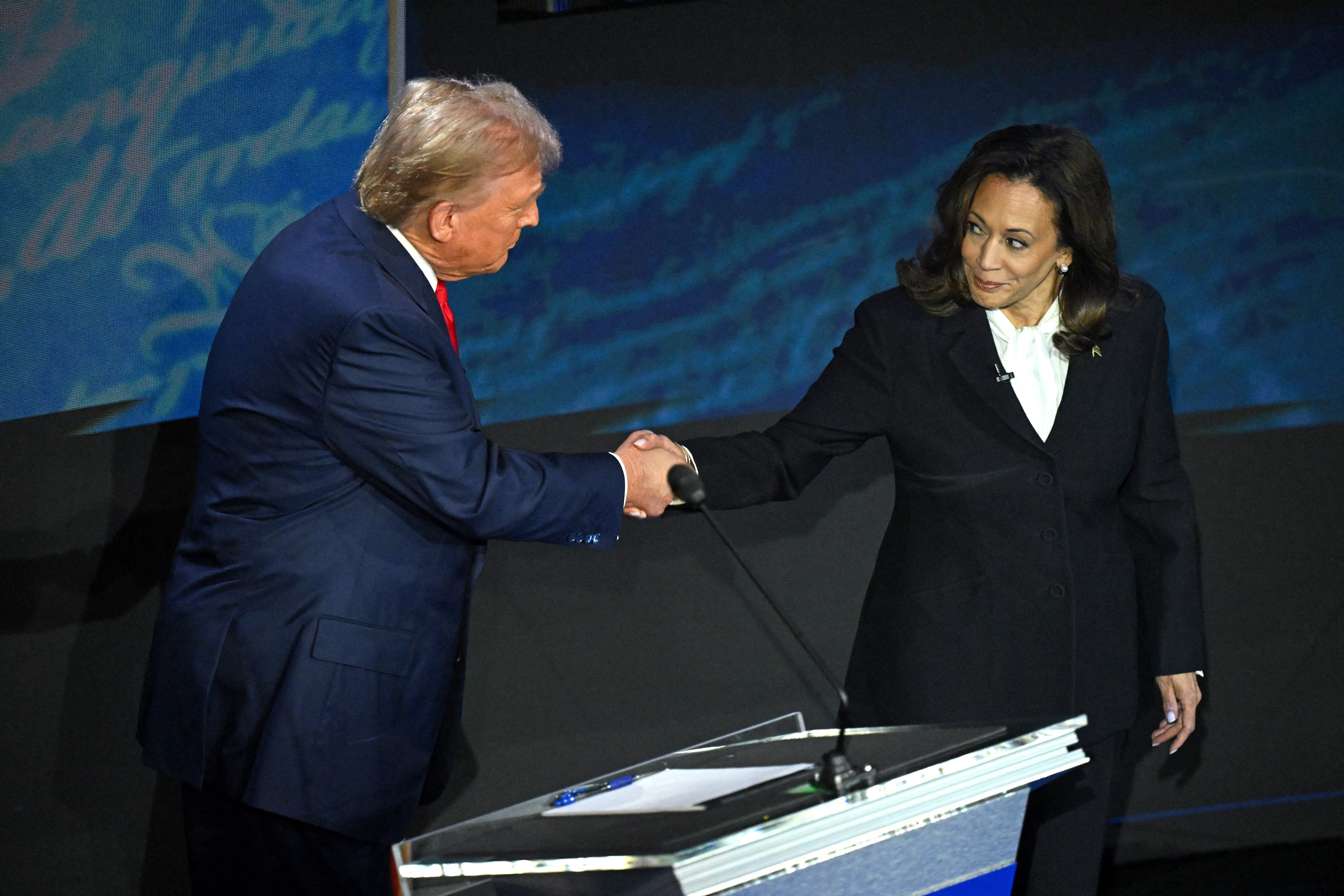 Donald Trump in a blue suit with a red tie shakes hands with Kamala Harris in a black suit with a white blouse in front of a podium.