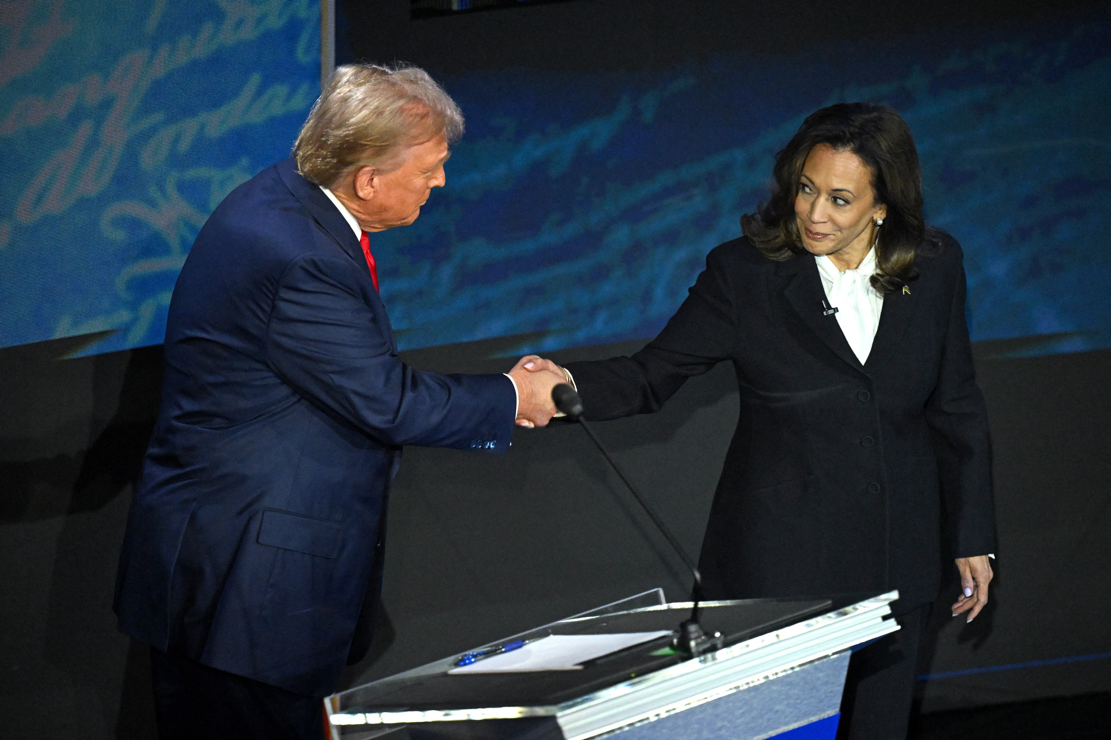 Donald Trump in a blue suit with a red tie shakes hands with Kamala Harris in a black suit with a white blouse in front of a podium.