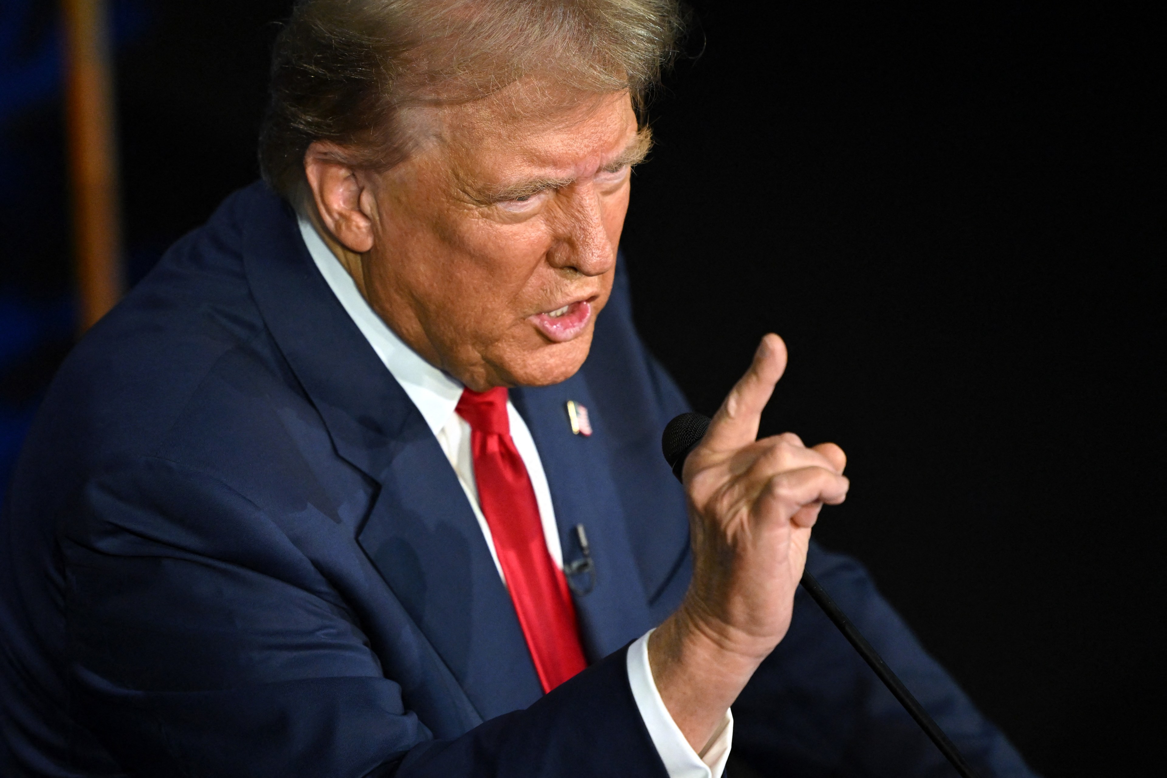 Donald Trump in a dark blue suit and red tie gestures with his index finger raised, seemingly in mid-speech. He holds a microphone close to his mouth.