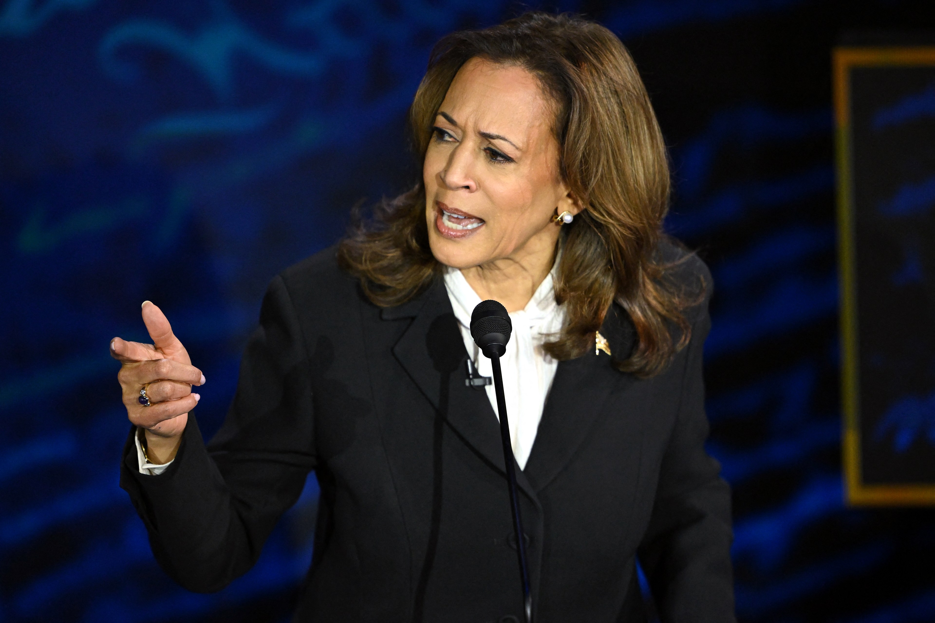 Kamala Harris in a black suit and white blouse speaks passionately into a microphone with her right hand gesturing and a determined expression on her face against a dark background.