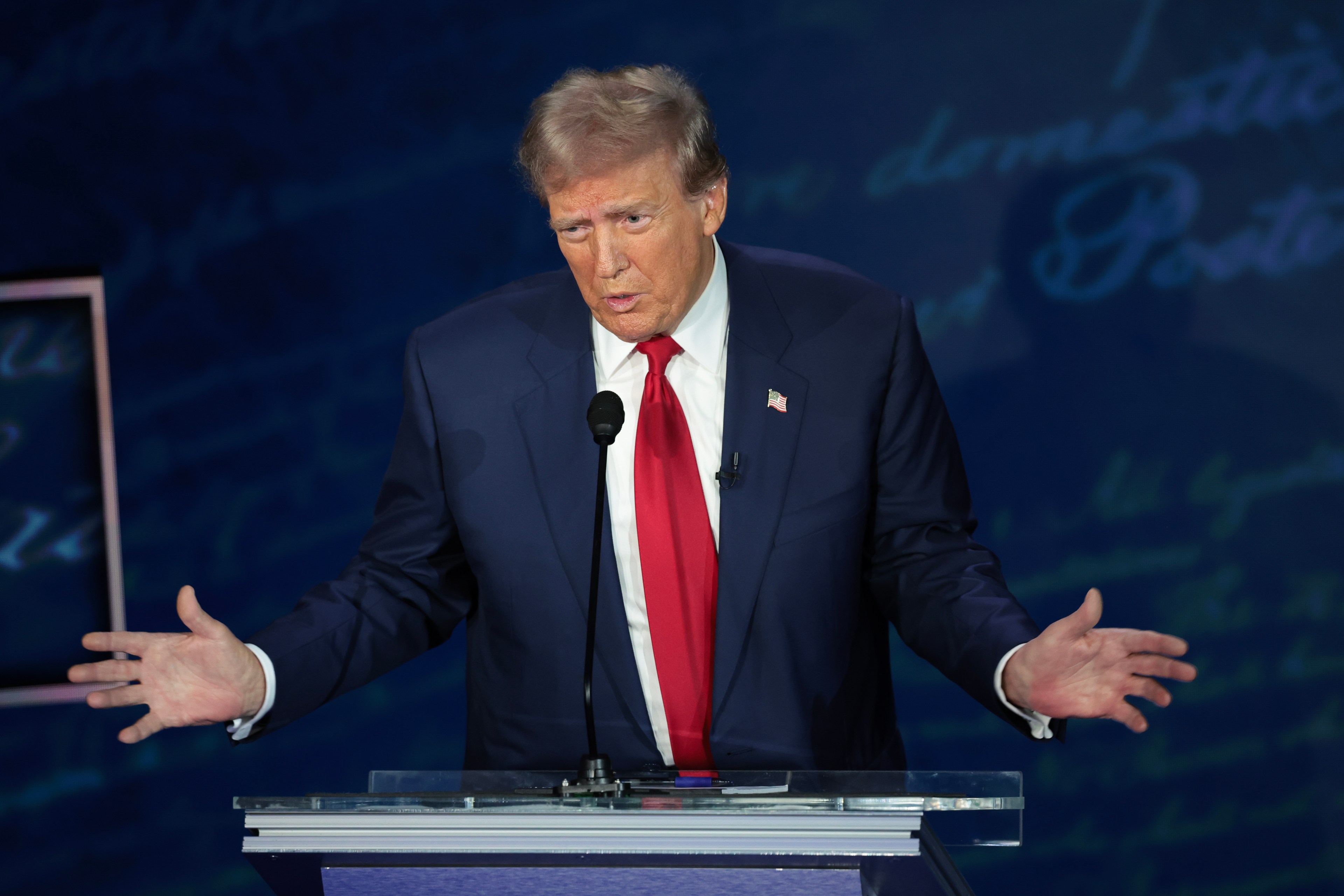 A man in a suit and red tie stands at a clear podium with a microphone, gesturing with open hands. Behind him is a blurred background with cursive writing.