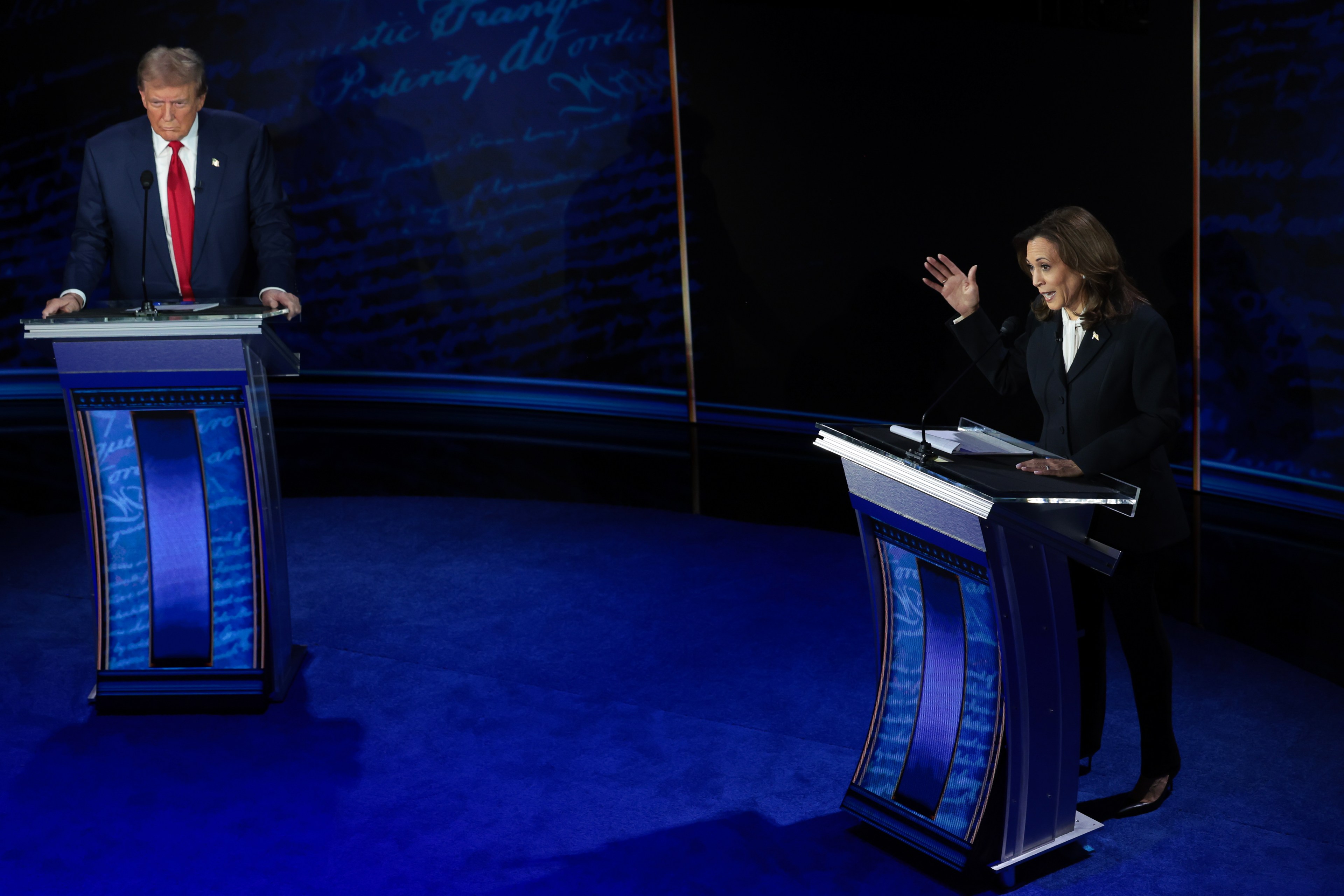 Two people stand behind separate blue-lit podiums on a debate stage; one is speaking animatedly with a hand raised, while the other looks down, showing a serious expression.